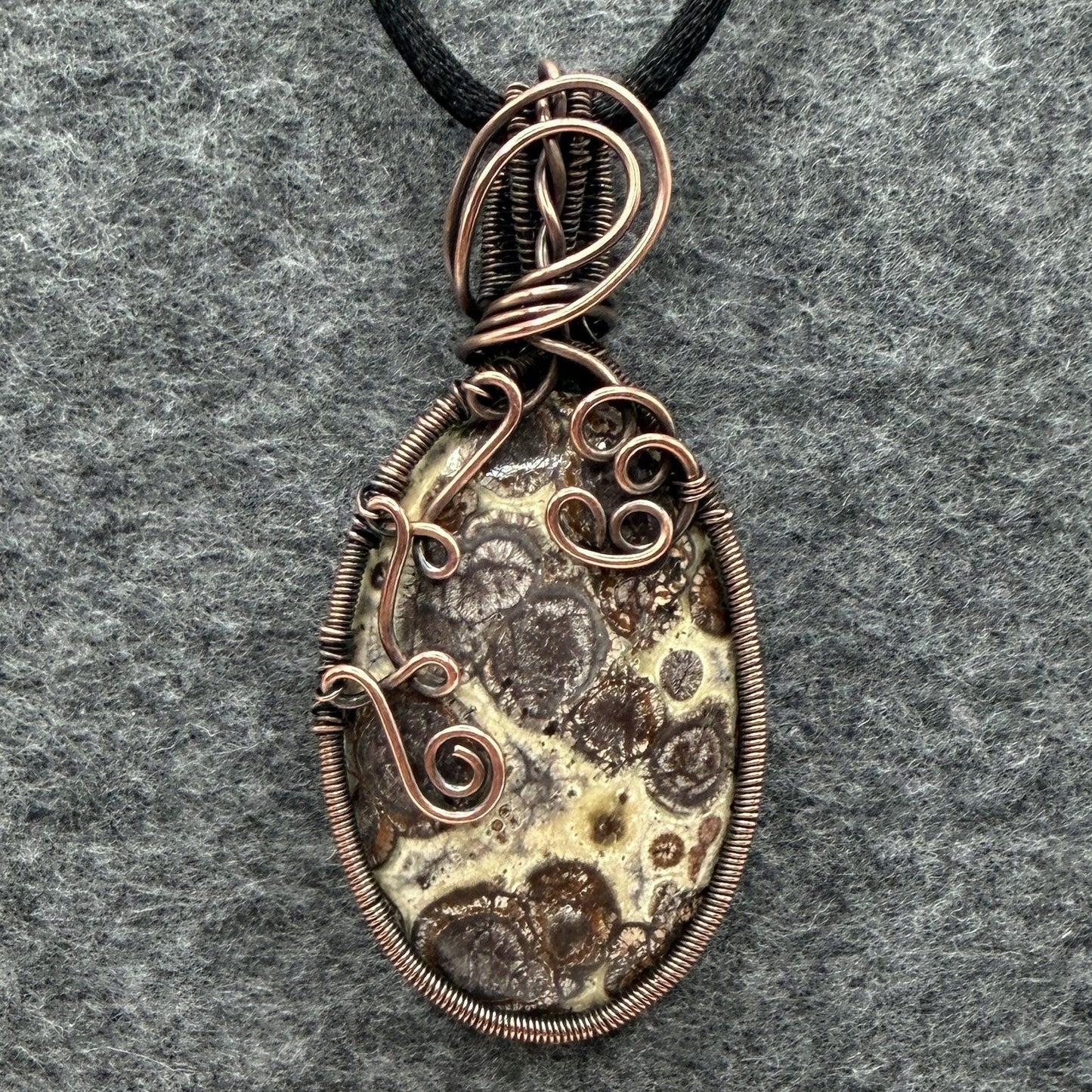 Front view of a handmade oval pendant necklace featuring a natural brown and cream asteroid jasper stone, wrapped in antiqued copper wire with decorative swirls, hanging from a black cord on a textured gray background.