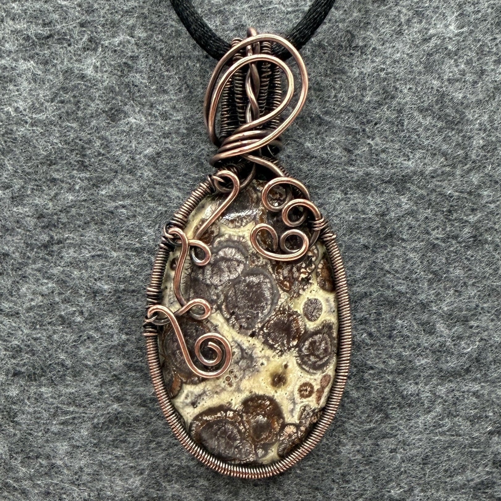 Front view of a handmade oval pendant necklace featuring a natural brown and cream asteroid jasper stone, wrapped in antiqued copper wire with decorative swirls, hanging from a black cord on a textured gray background.