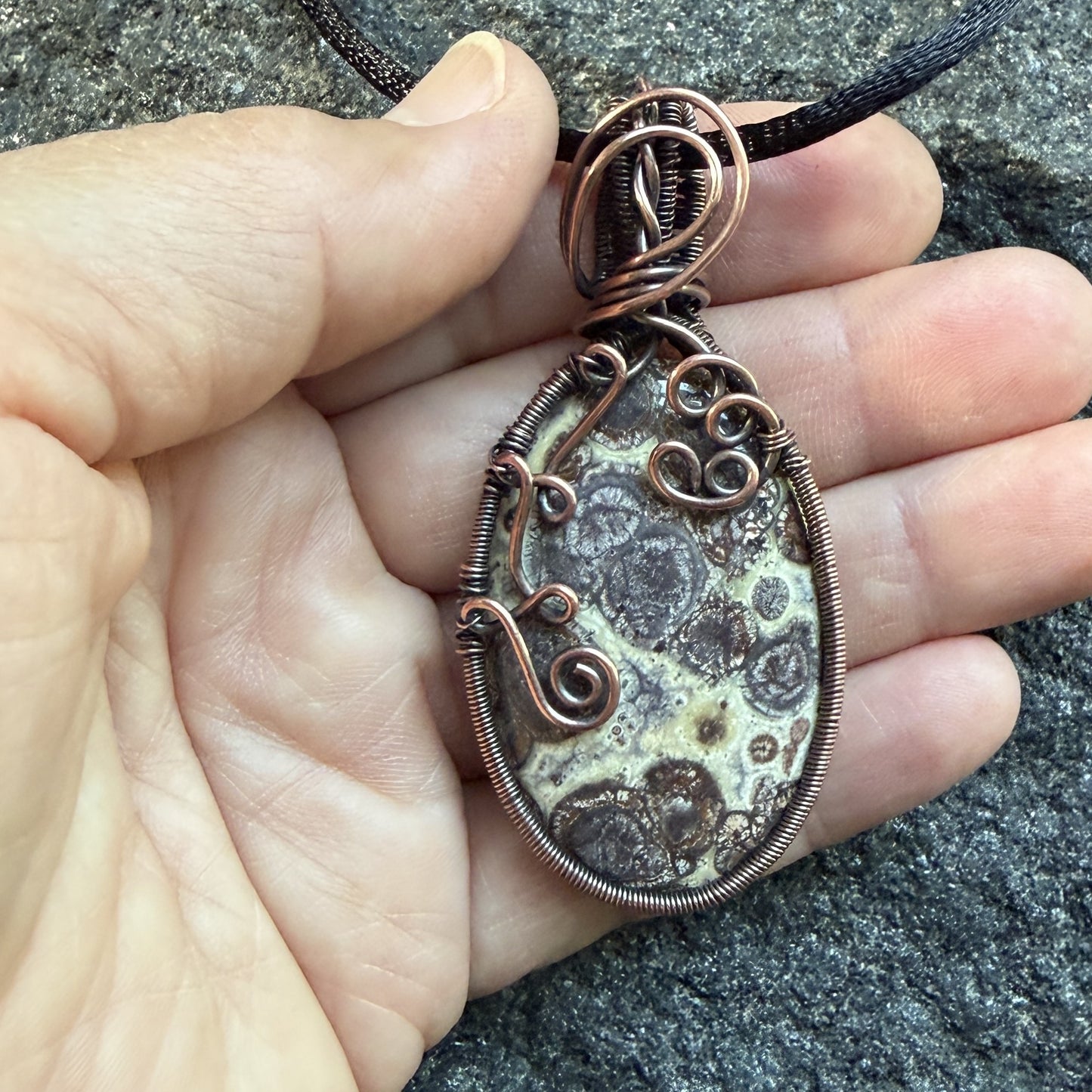 Front view of a woven-framed handmade oval pendant necklace featuring a natural brown and cream asteroid jasper stone, wrapped in antiqued copper wire with decorative swirls, hanging from a black cord, held in the artist's hand.