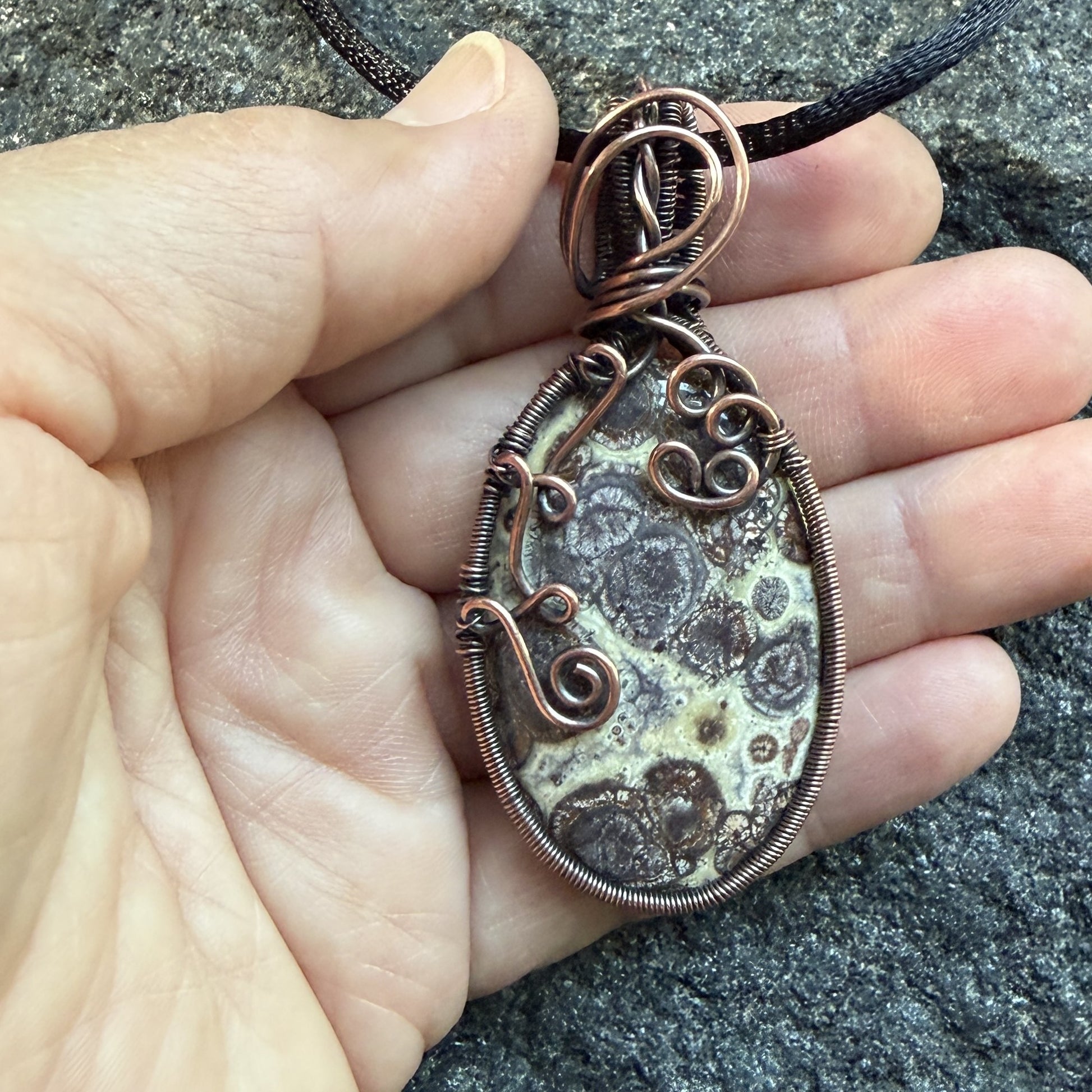 Front view of a woven-framed handmade oval pendant necklace featuring a natural brown and cream asteroid jasper stone, wrapped in antiqued copper wire with decorative swirls, hanging from a black cord, held in the artist's hand.