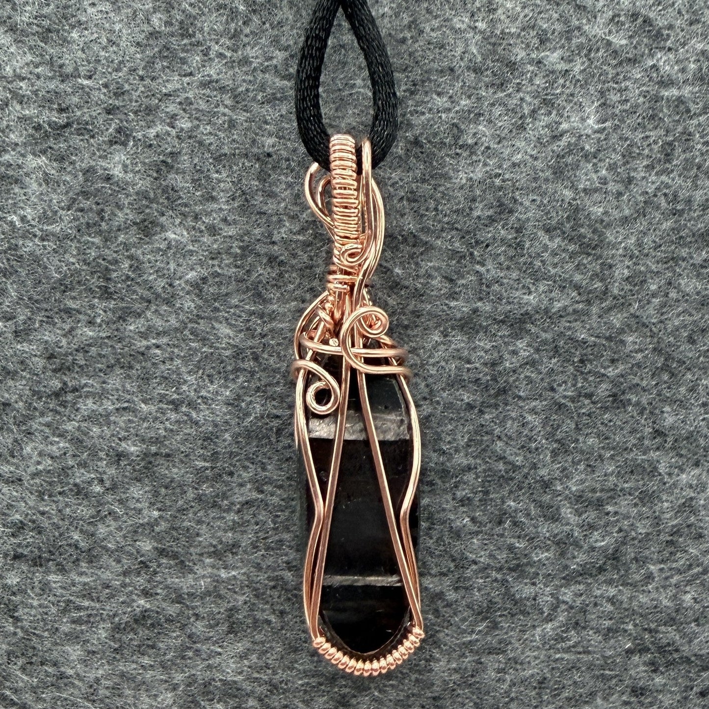 Back view of a handmade pendant necklace featuring a polished black and silver hypersthene stone wrapped in copper wire, suspended from a black cord against a textured gray felt background.