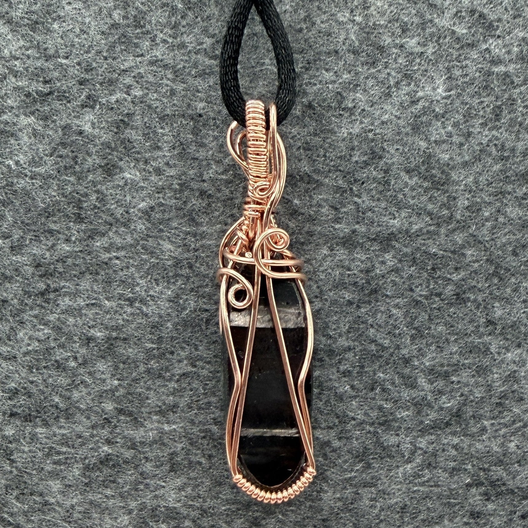 Back view of a handmade pendant necklace featuring a polished black and silver hypersthene stone wrapped in copper wire, suspended from a black cord against a textured gray felt background.