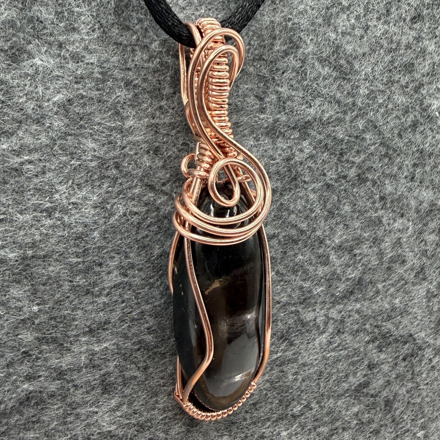 Detail view of a handmade pendant necklace featuring a polished black and silver hypersthene stone wrapped in copper wire, suspended from a black cord against a textured gray felt background.