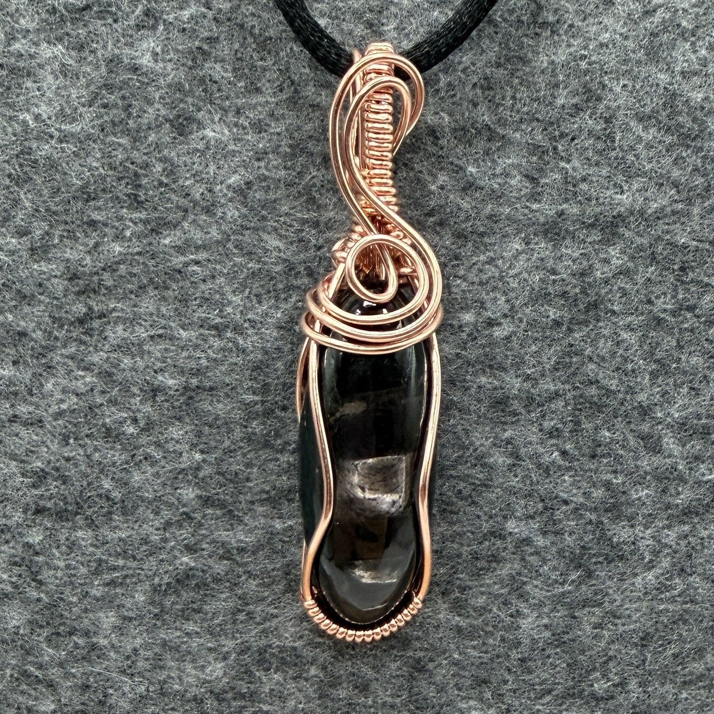 Front view of a handmade pendant necklace featuring a polished black and silver hypersthene stone wrapped in copper wire, suspended from a black cord against a textured gray felt background.