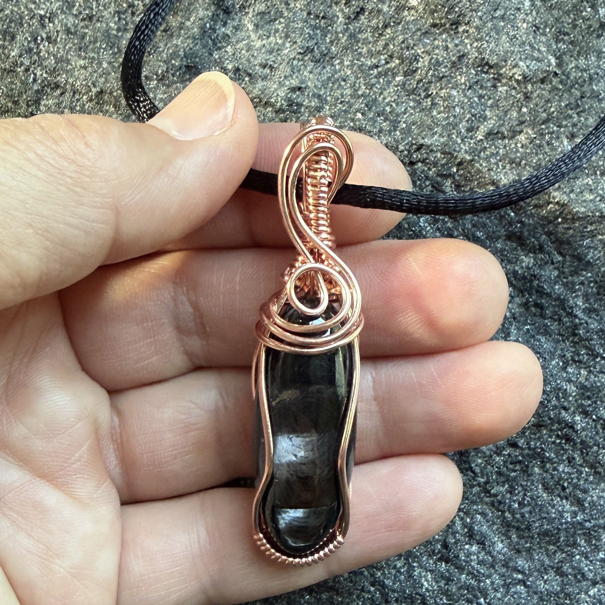 Front view of a handmade pendant necklace featuring a polished black and silver hypersthene stone wrapped in copper wire, suspended from a black cord, held in the artist's hand.