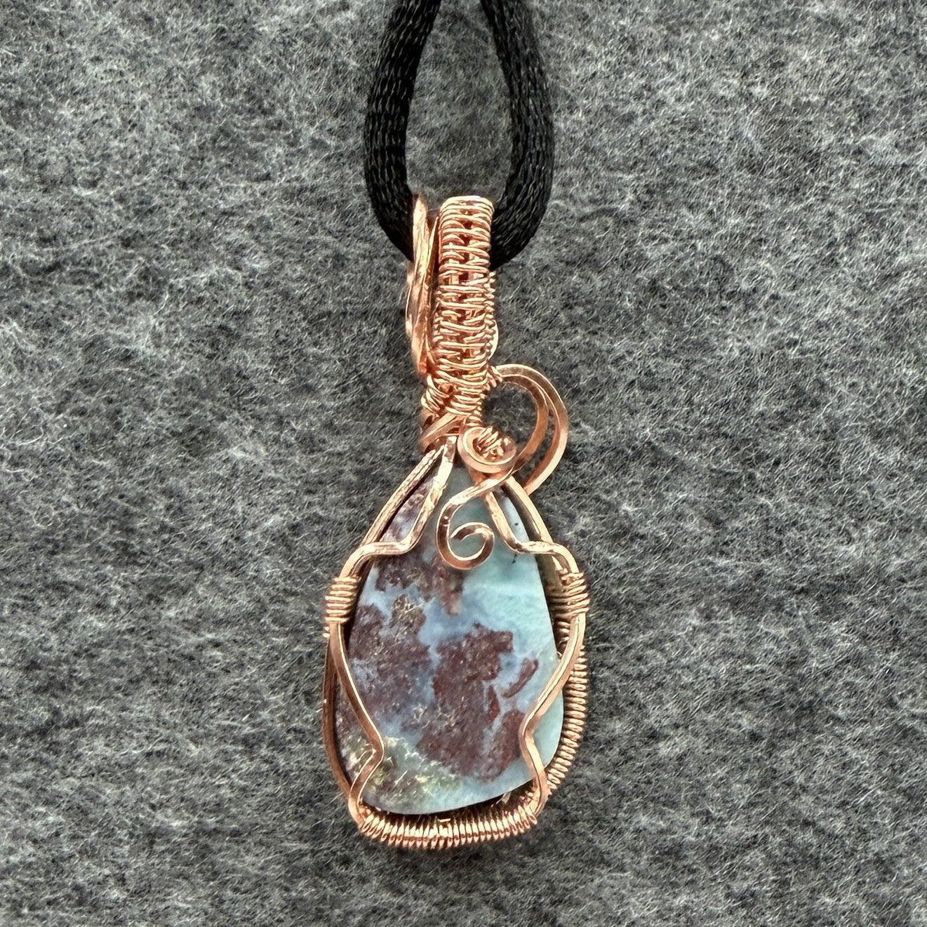 Back view of a teardrop-shaped wire-wrapped pendant necklace featuring a blue and brown marbled larimar stone, suspended from a black cord against a textured gray background.
