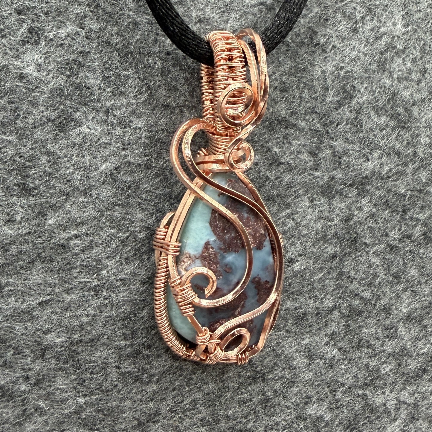 Detail view of a teardrop-shaped wire-wrapped pendant necklace featuring a blue and brown marbled larimar stone, suspended from a black cord against a textured gray background.