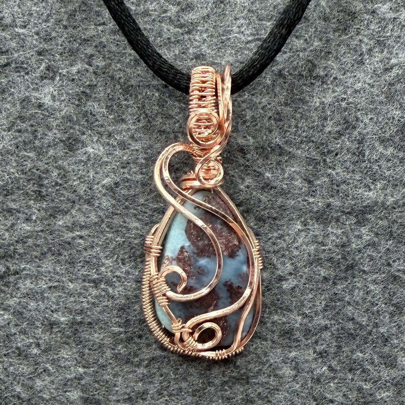 Front view of a teardrop-shaped wire-wrapped pendant necklace featuring a blue and brown marbled larimar stone, suspended from a black cord against a textured gray background.