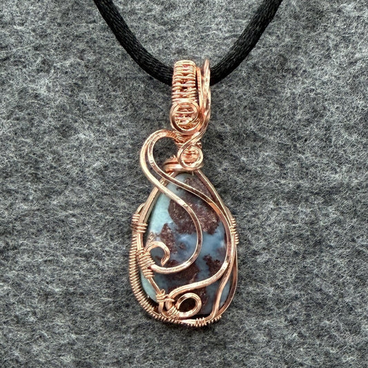 Front view of a teardrop-shaped wire-wrapped pendant necklace featuring a blue and brown marbled larimar stone, suspended from a black cord against a textured gray background.