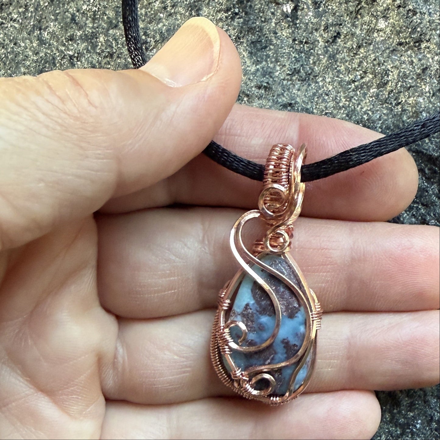 Front view of a teardrop-shaped wire-wrapped pendant necklace featuring a blue and brown marbled larimar stone, suspended from a black cord, held in the artist's hand.