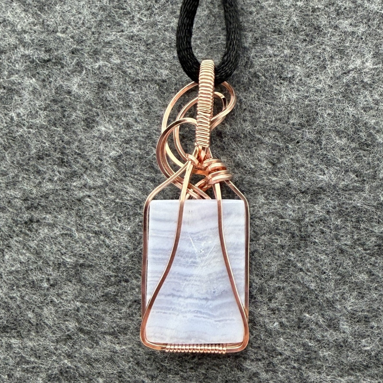 Back view of a rectangular pendant necklace featuring a blue lace agate stone wrapped in copper wire, suspended from a black cord, set against a textured gray background.