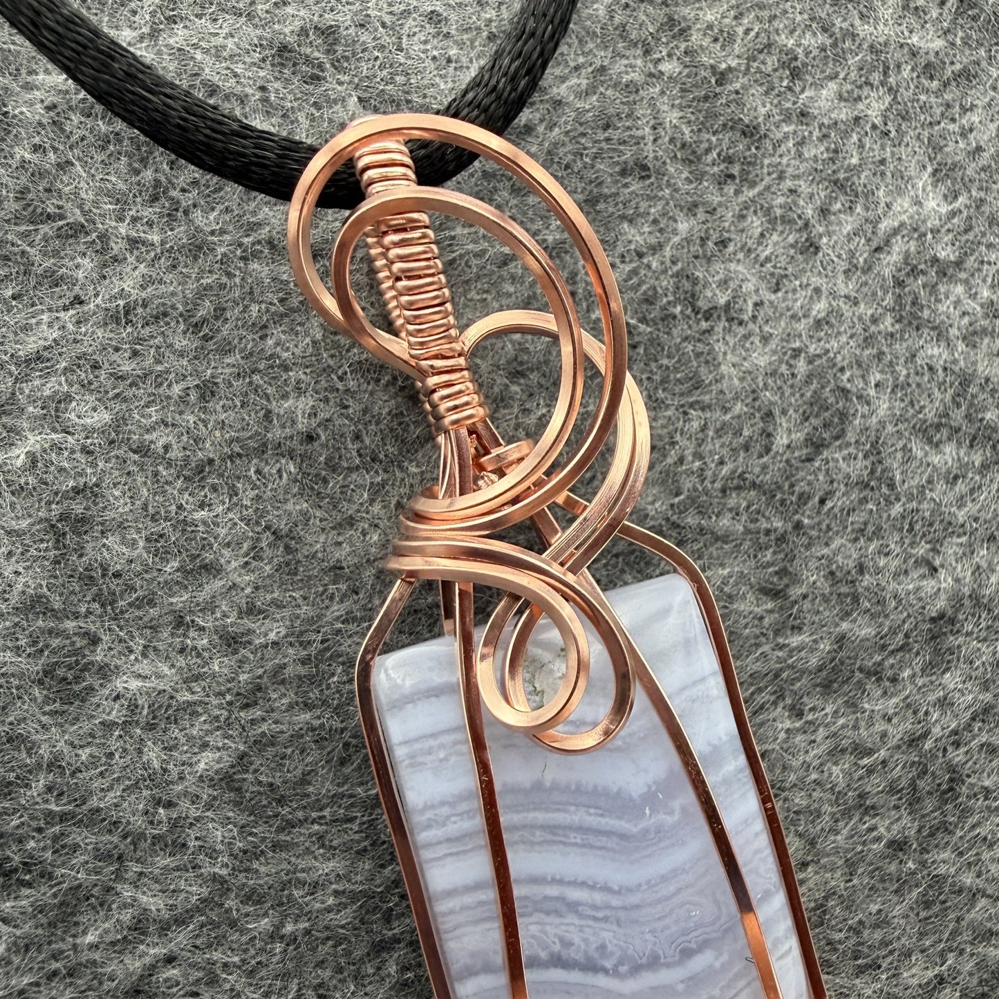 Detail view of a rectangular pendant necklace featuring a blue lace agate stone wrapped in copper wire, suspended from a black cord, set against a textured gray background.
