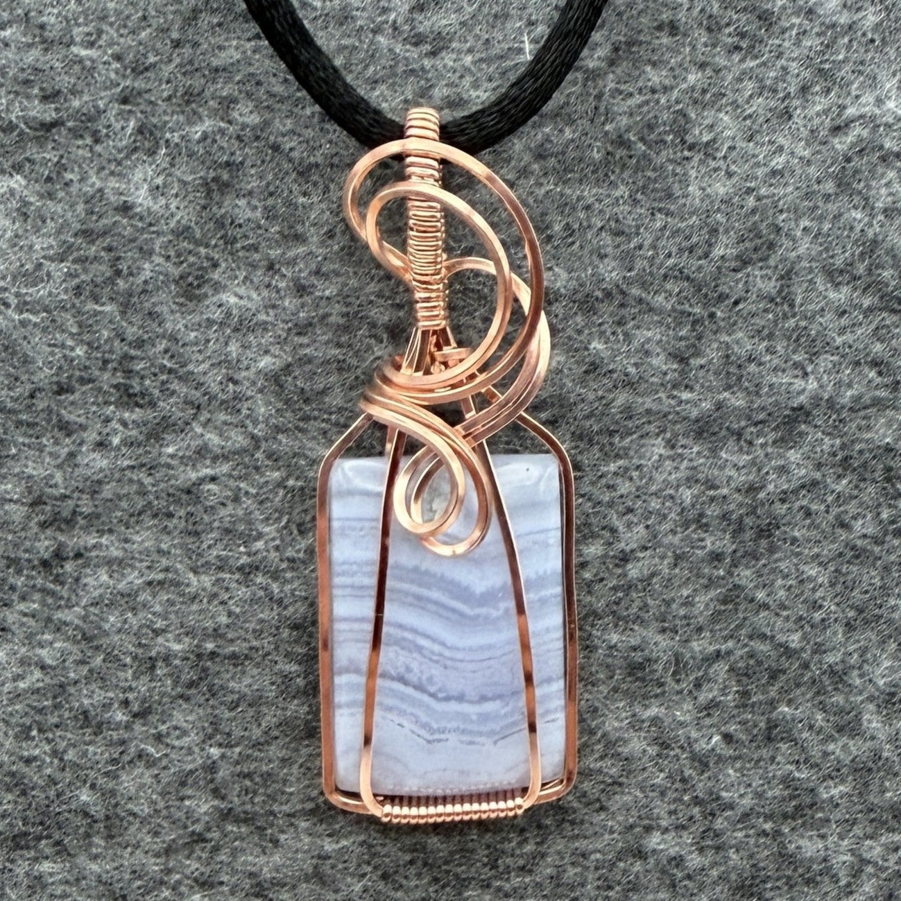 Front view of a rectangular pendant necklace featuring a blue lace agate stone wrapped in copper wire, suspended from a black cord, set against a textured gray background.