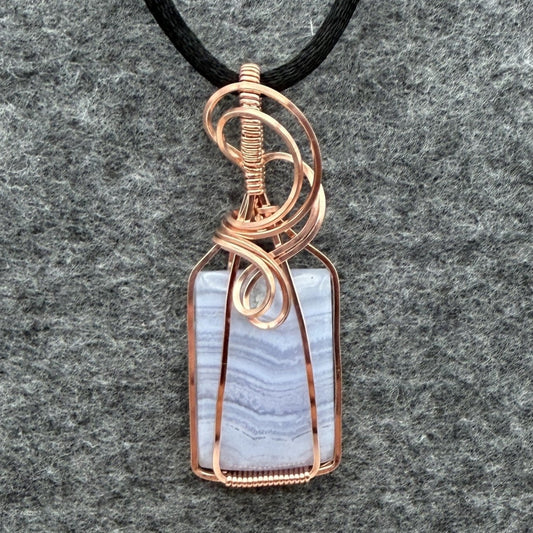 Front view of a rectangular pendant necklace featuring a blue lace agate stone wrapped in copper wire, suspended from a black cord, set against a textured gray background.