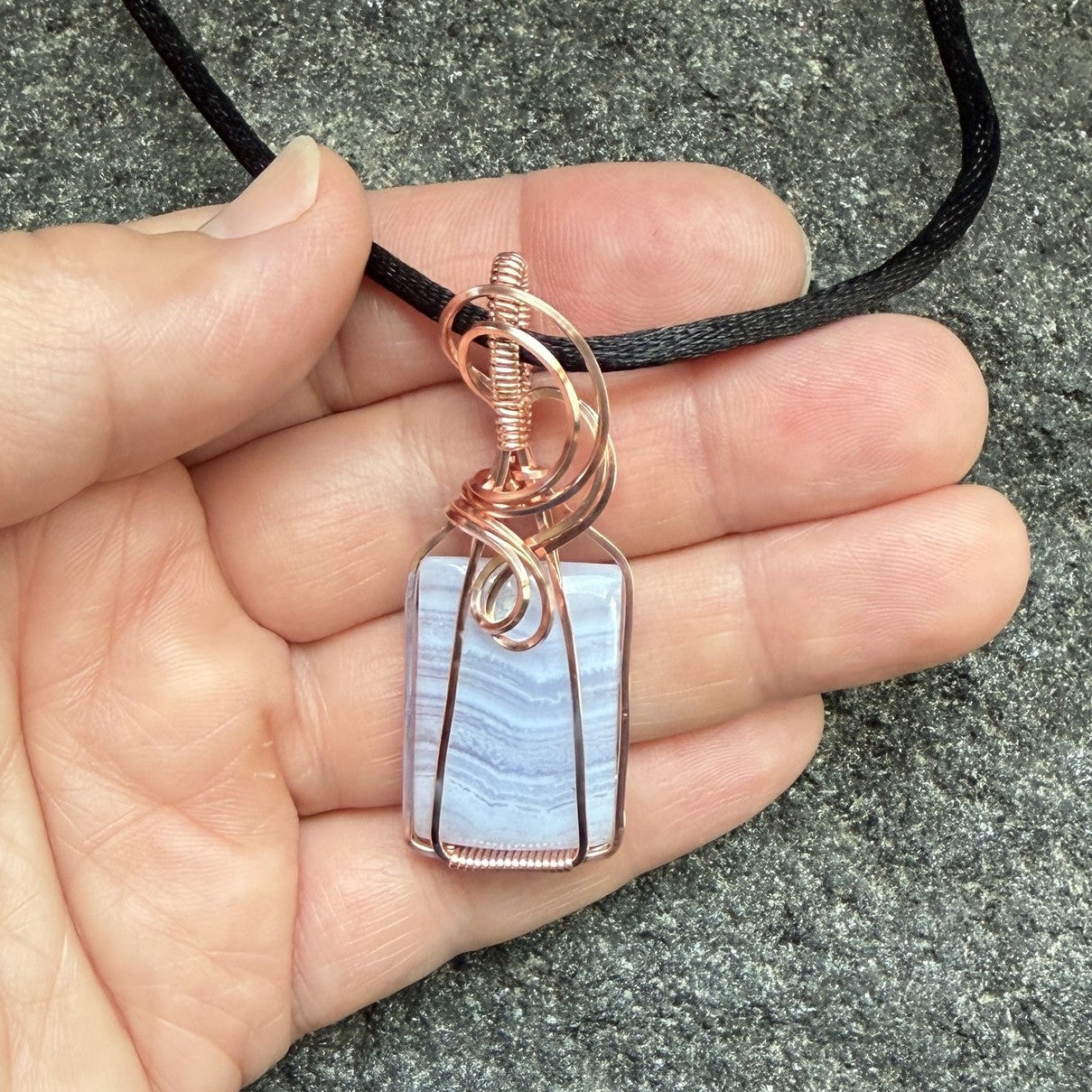 Front view of a rectangular pendant necklace featuring a blue lace agate stone wrapped in copper wire, suspended from a black cord, held in the artist's hand.