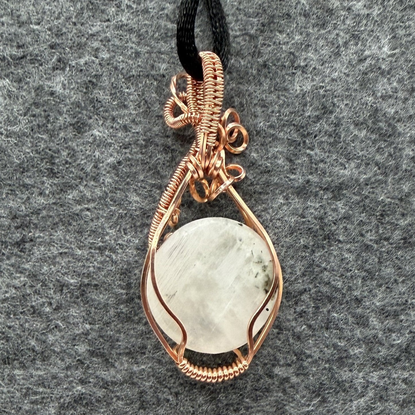 Back view of a handmade teardrop pendant necklace featuring a round, milky white moonstone wrapped in textured copper wire, suspended from a simple black cord, set against a dark gray felt background.