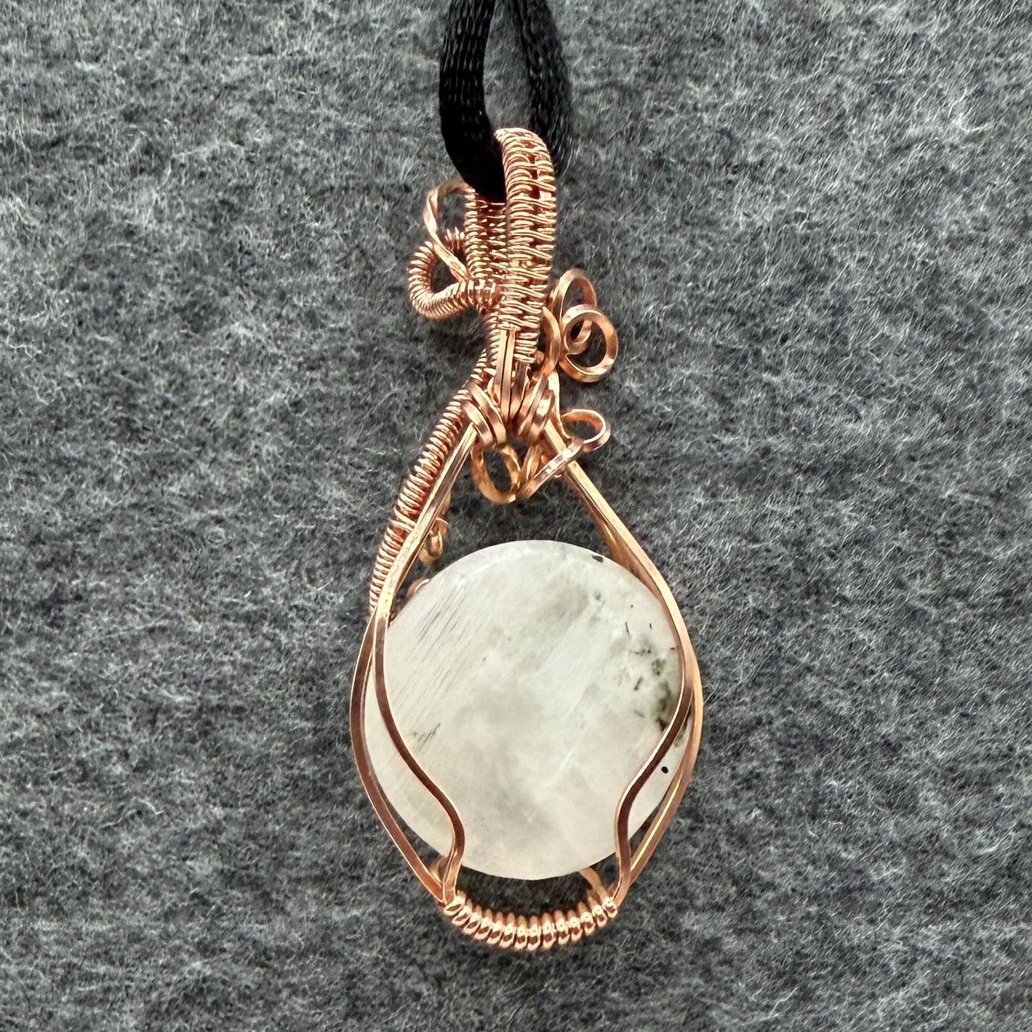 Back view of a handmade teardrop pendant necklace featuring a round, milky white moonstone wrapped in textured copper wire, suspended from a simple black cord, set against a dark gray felt background.
