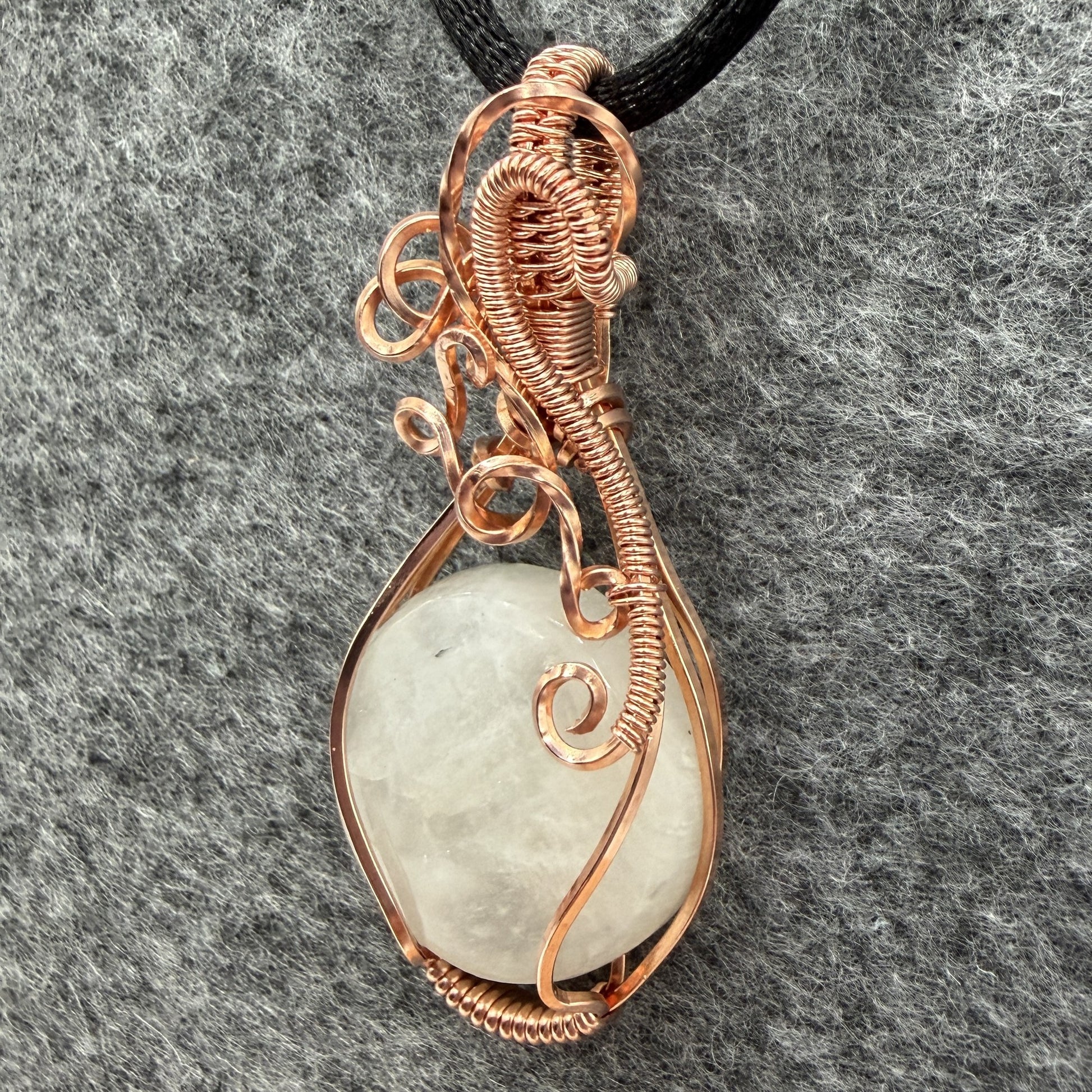 Detail view of a handmade teardrop pendant necklace featuring a round, milky white moonstone wrapped in textured copper wire, suspended from a simple black cord, set against a dark gray felt background.