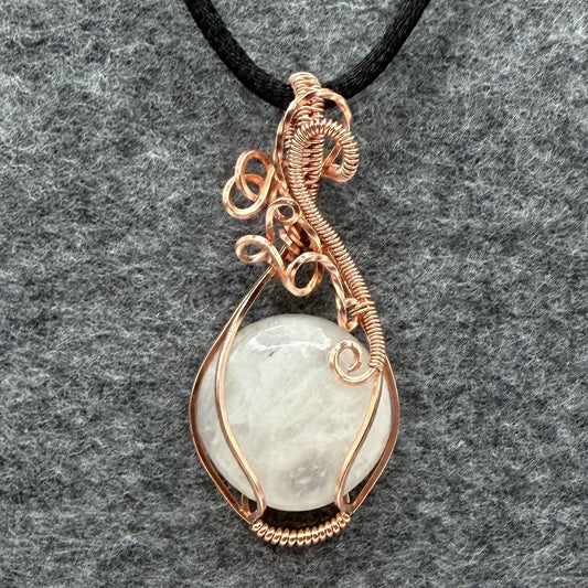 Front view of a handmade teardrop pendant necklace featuring a round, milky white moonstone wrapped in textured copper wire, suspended from a simple black cord, set against a dark gray felt background.