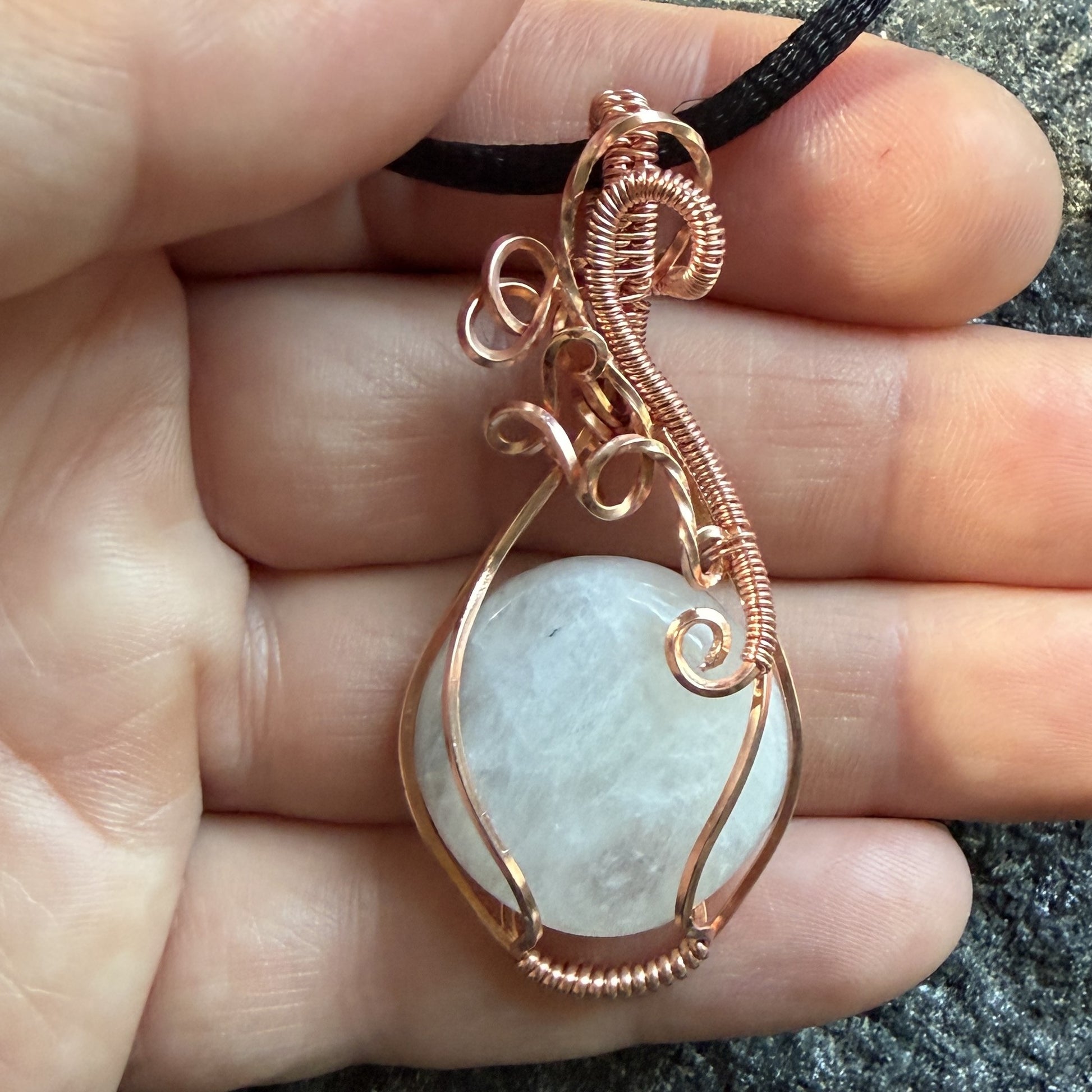 Front view of a handmade teardrop pendant necklace featuring a round, milky white moonstone wrapped in textured copper wire, suspended from a simple black cord, held in the artist's hand.