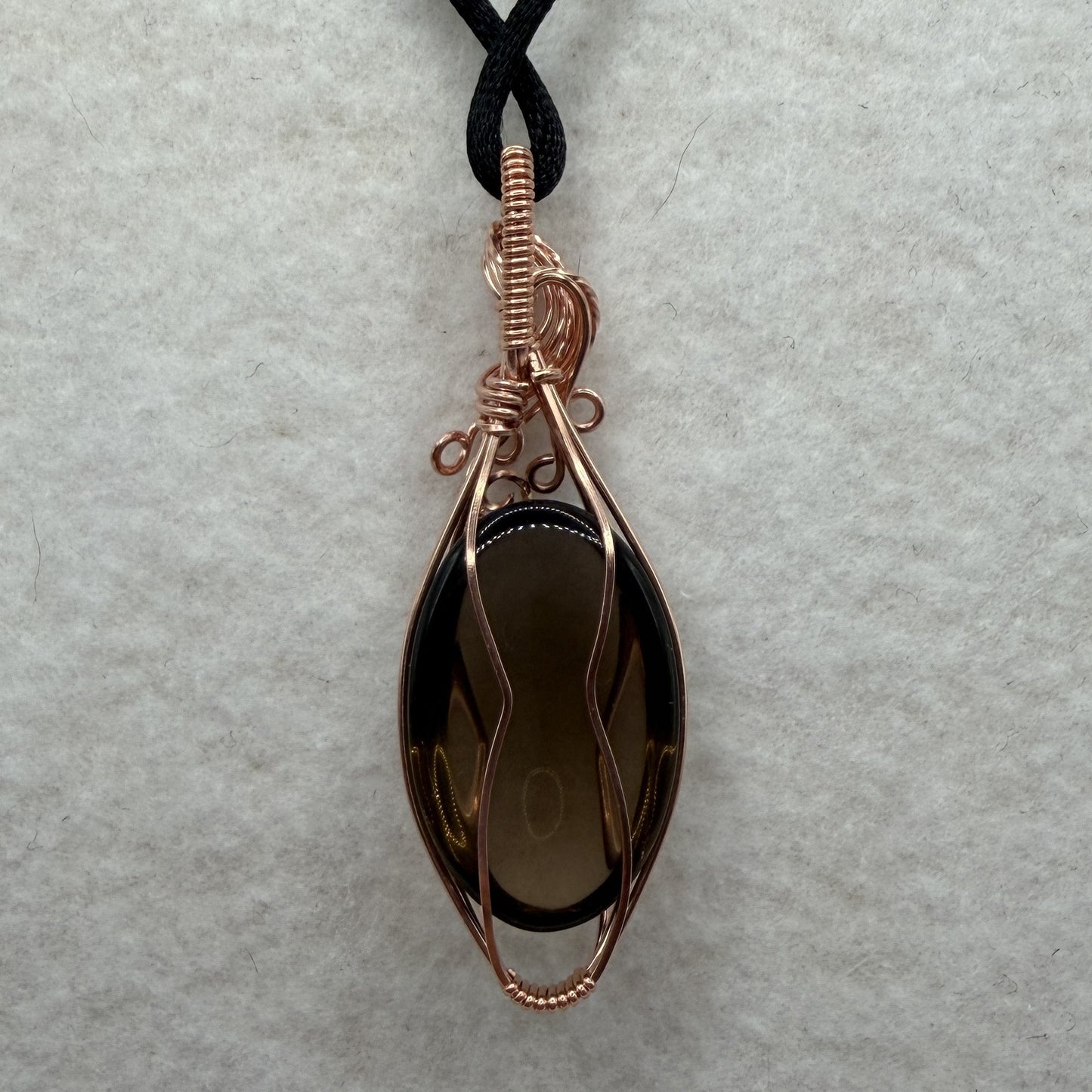 Back view of a teardrop-shaped dark brown smoky quartz stone pendant wrapped in copper wire, hanging from a black cord on a textured white background.