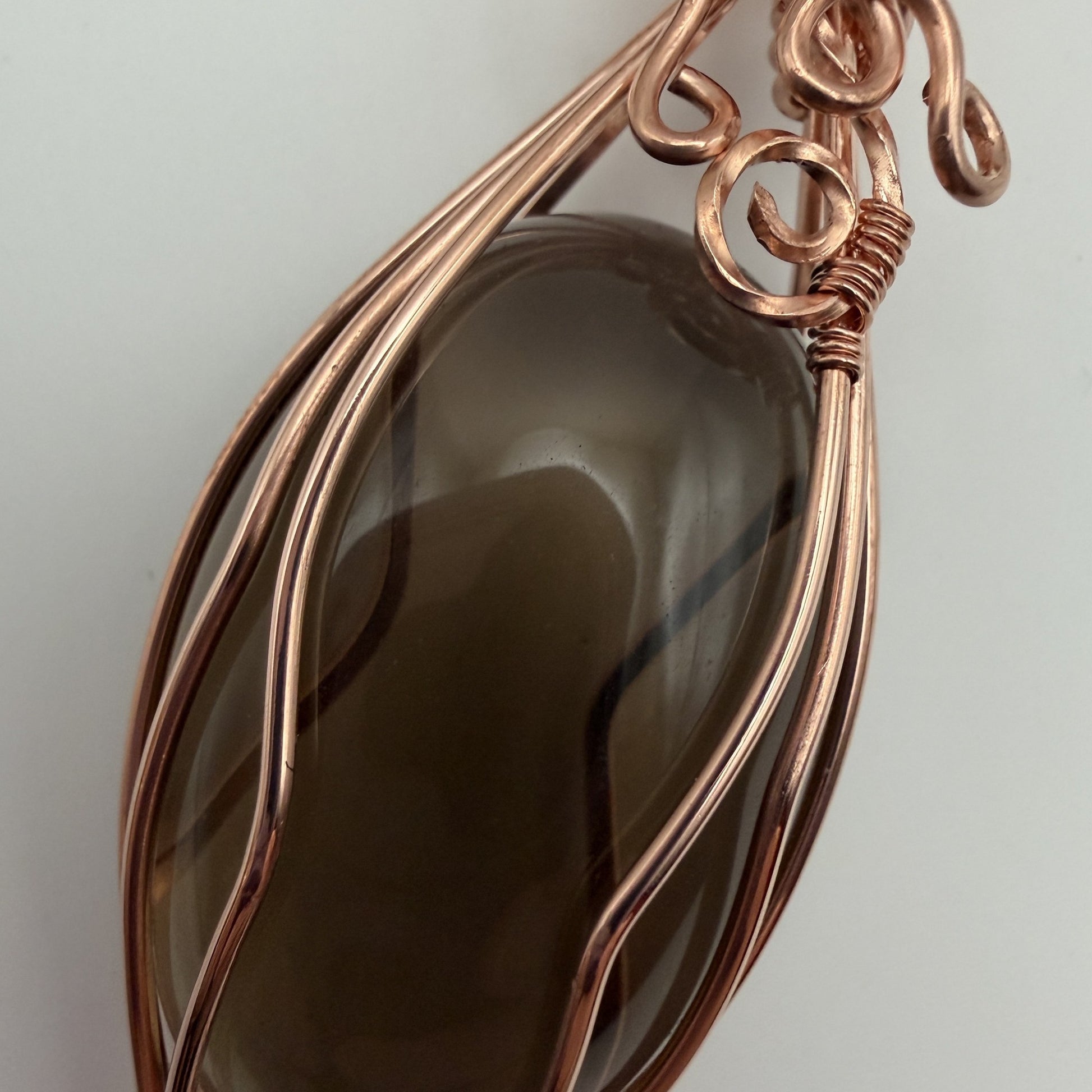 Detail view of a teardrop-shaped dark brown smoky quartz stone pendant wrapped in copper wire, hanging from a black cord on a textured white background.