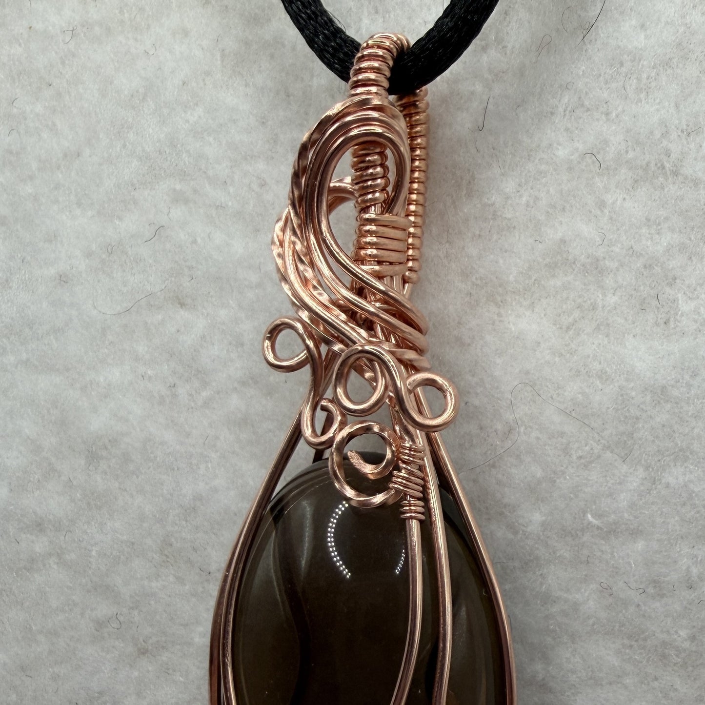 Detail view of a teardrop-shaped dark brown smoky quartz stone pendant wrapped in copper wire, hanging from a black cord on a textured white background.