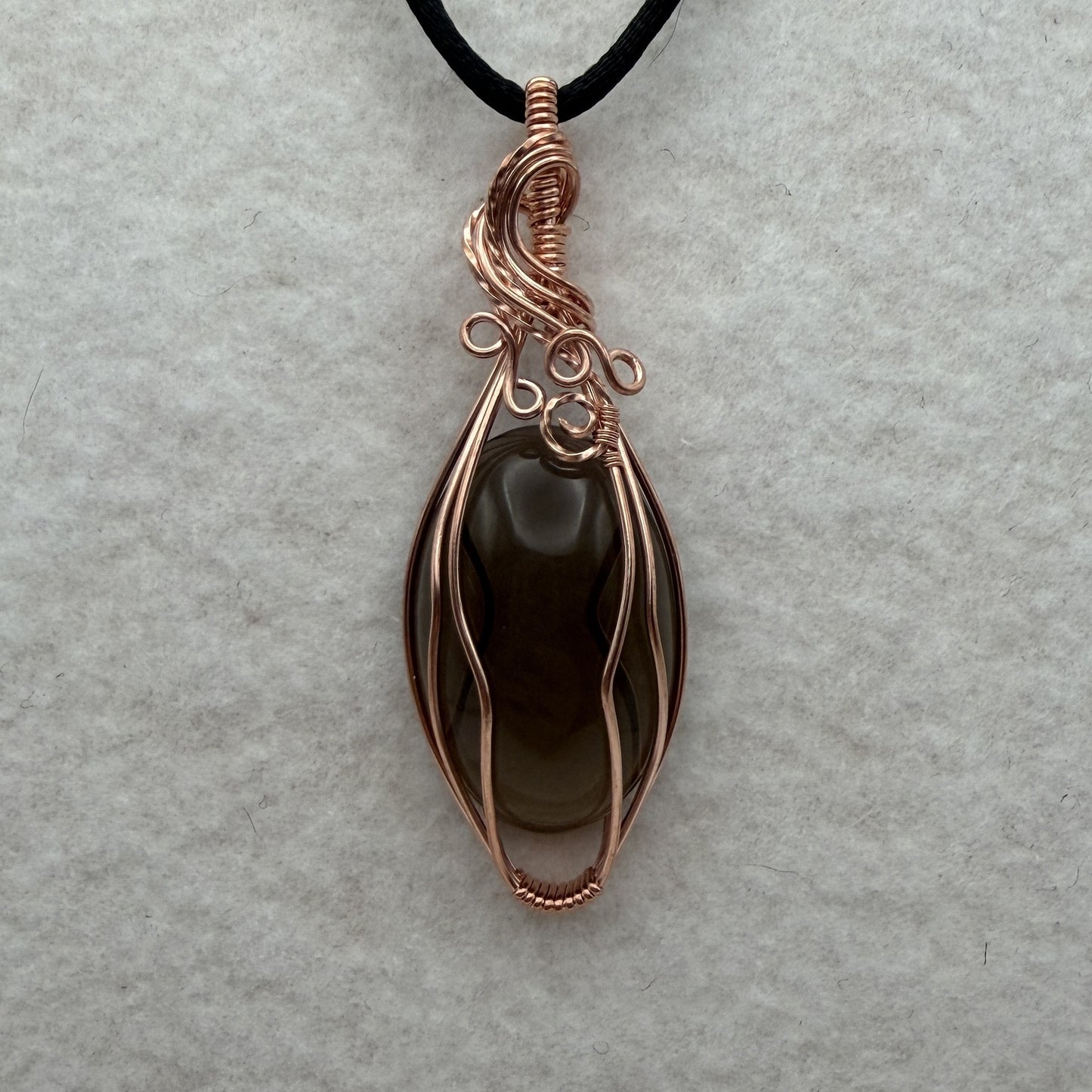 Front view of a teardrop-shaped dark brown smoky quartz stone pendant wrapped in copper wire, hanging from a black cord on a textured white background.
