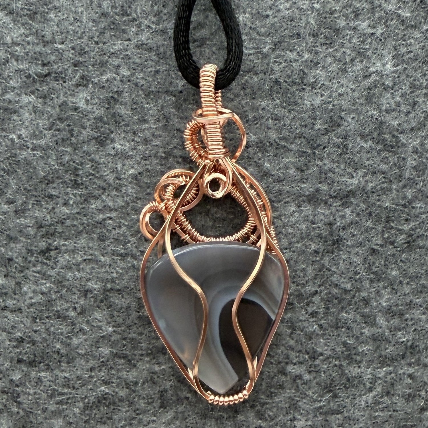 Back view of a handmade wire-wrapped pendant necklace featuring a botswana agate stone in a copper wire setting, suspended from a black cord against a textured gray background.