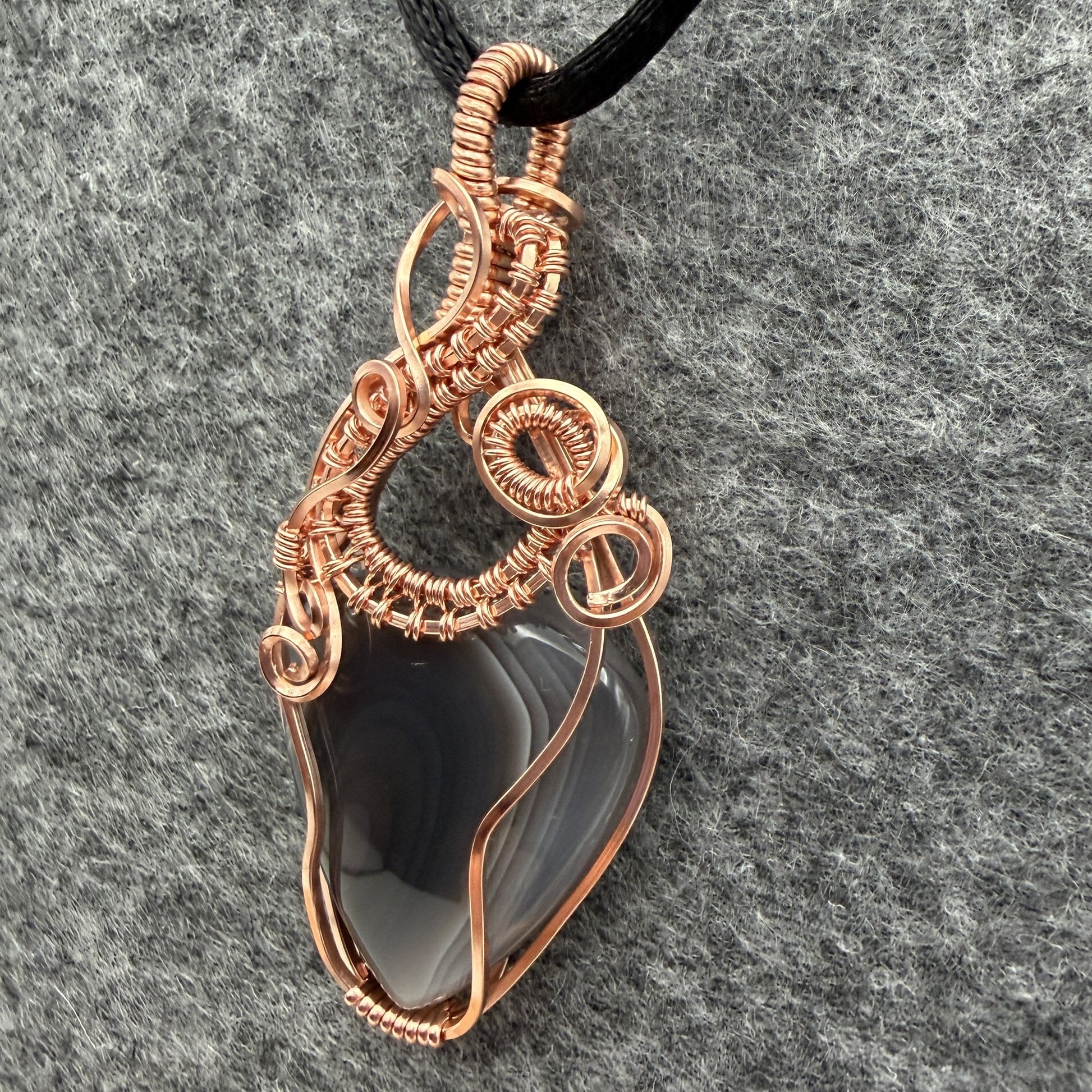 Detail view of a handmade wire-wrapped pendant necklace featuring a botswana agate stone in a copper wire setting, suspended from a black cord against a textured gray background.