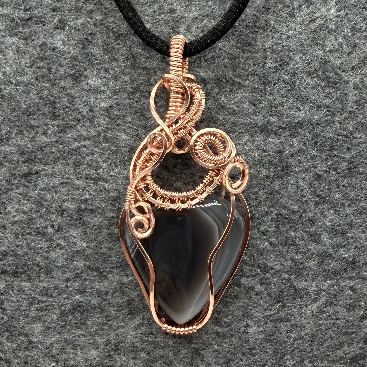 Front view of a handmade wire-wrapped pendant necklace featuring a botswana agate stone in a copper wire setting, suspended from a black cord against a textured gray background.