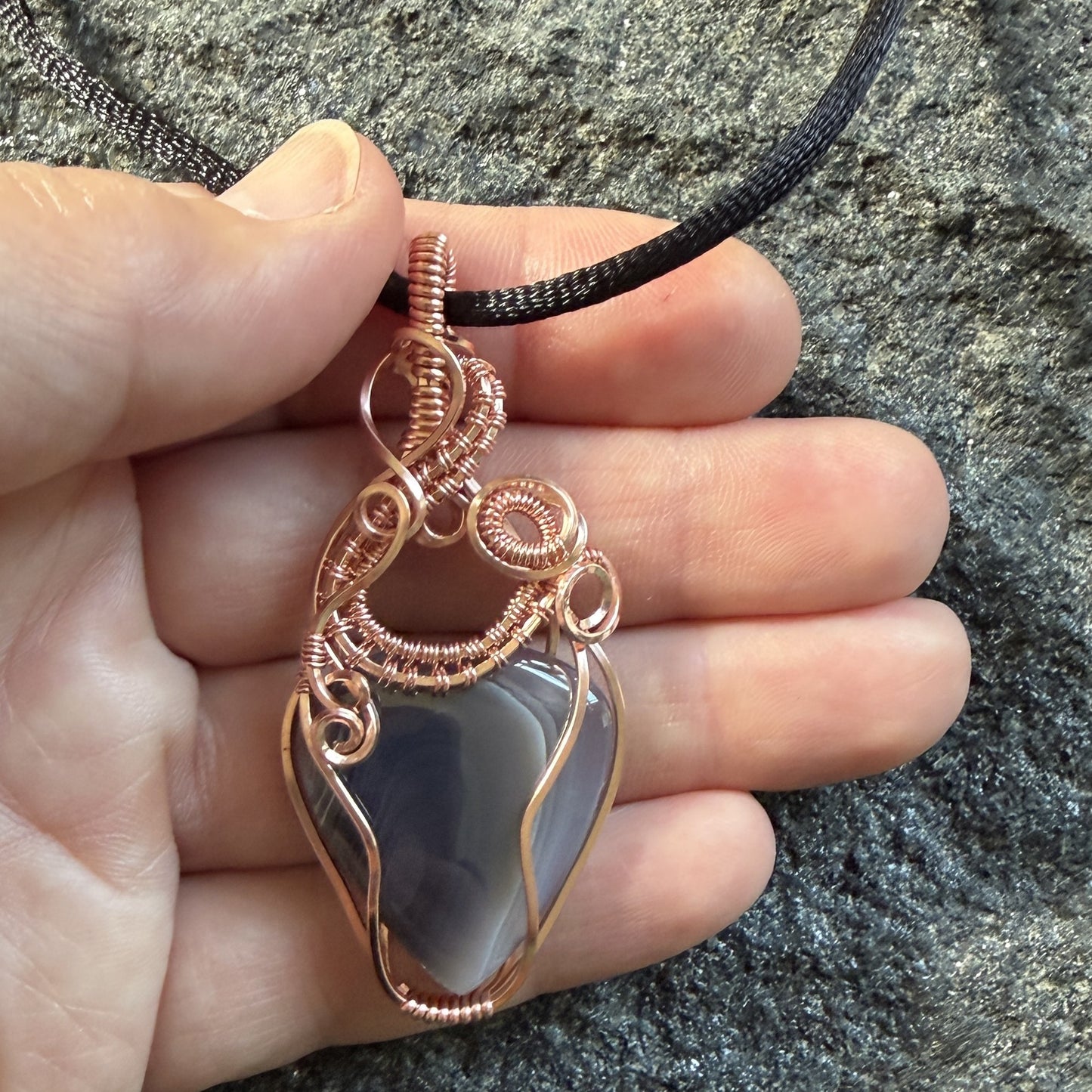 Front view of a handmade wire-wrapped pendant necklace featuring a botswana agate stone in a copper wire setting, suspended from a black cord, held in the artist's hand.