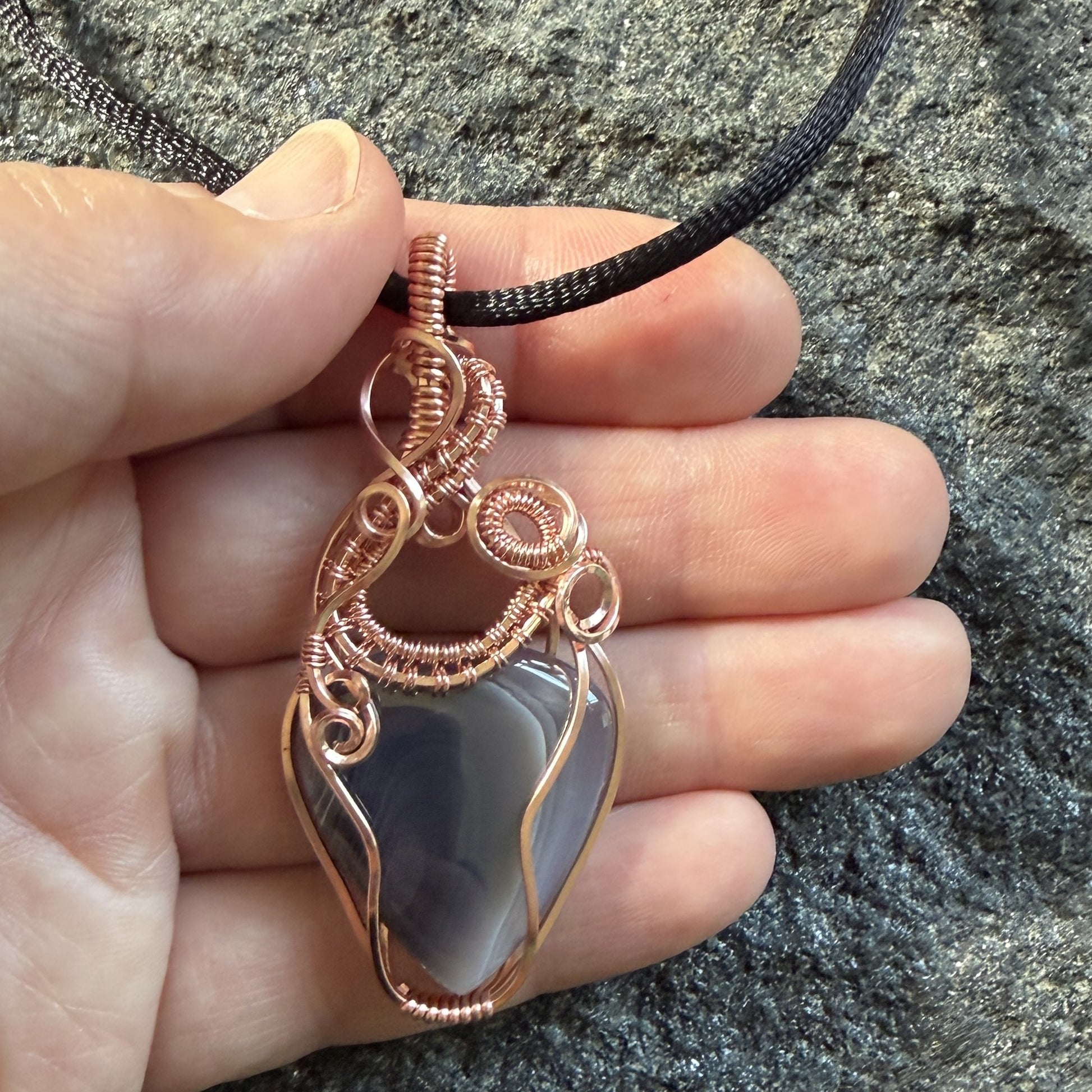 Front view of a handmade wire-wrapped pendant necklace featuring a botswana agate stone in a copper wire setting, suspended from a black cord, held in the artist's hand.