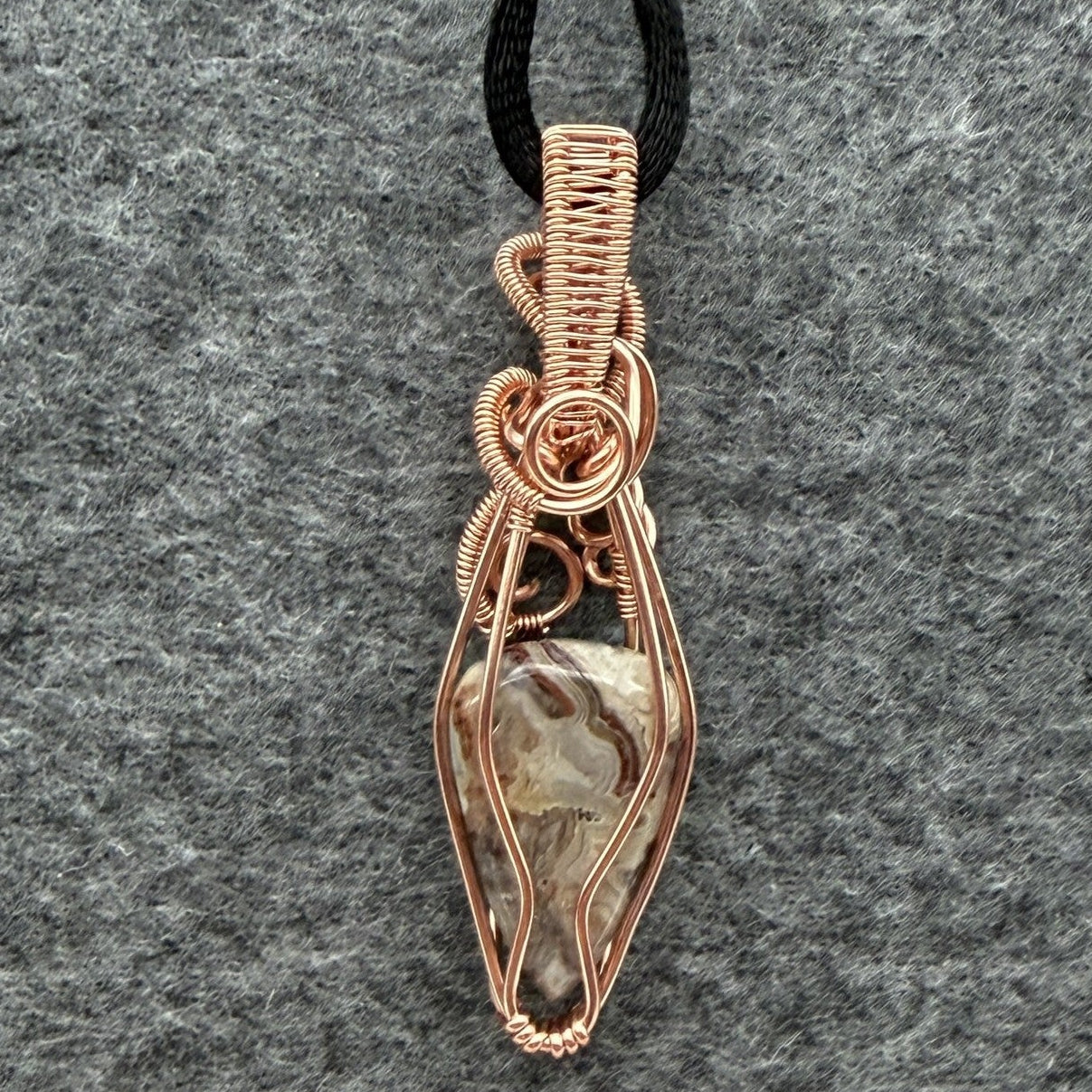 Back view of a handmade wire-wrapped pendant necklace featuring a crazy lace agate stone enclosed in copper wire, suspended from a black cord against a textured gray background.