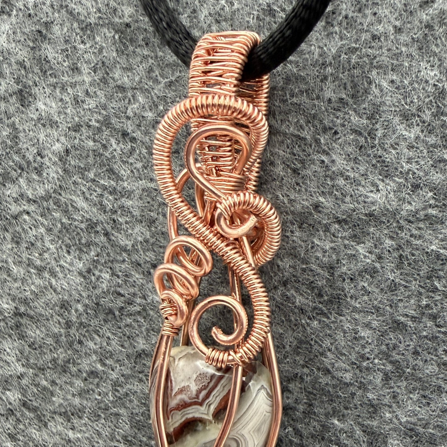 Detail view of a handmade wire-wrapped pendant necklace featuring a crazy lace agate stone enclosed in copper wire, suspended from a black cord against a textured gray background.