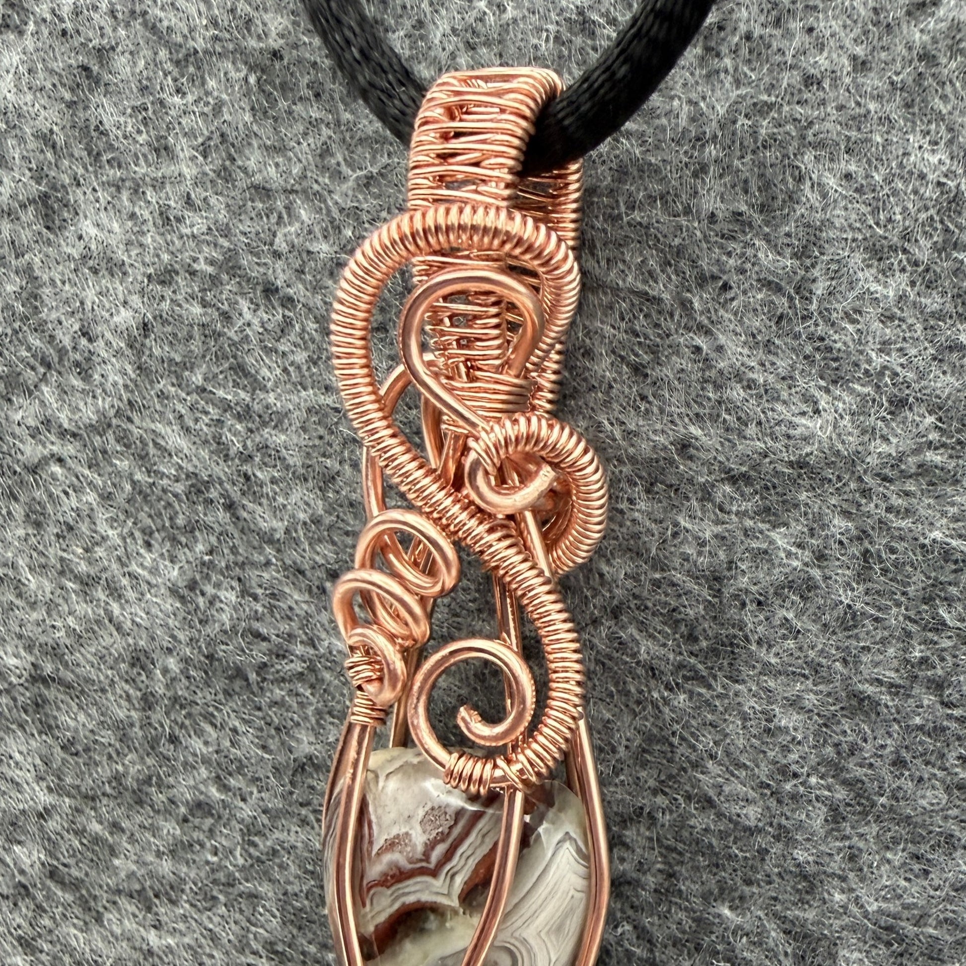 Detail view of a handmade wire-wrapped pendant necklace featuring a crazy lace agate stone enclosed in copper wire, suspended from a black cord against a textured gray background.