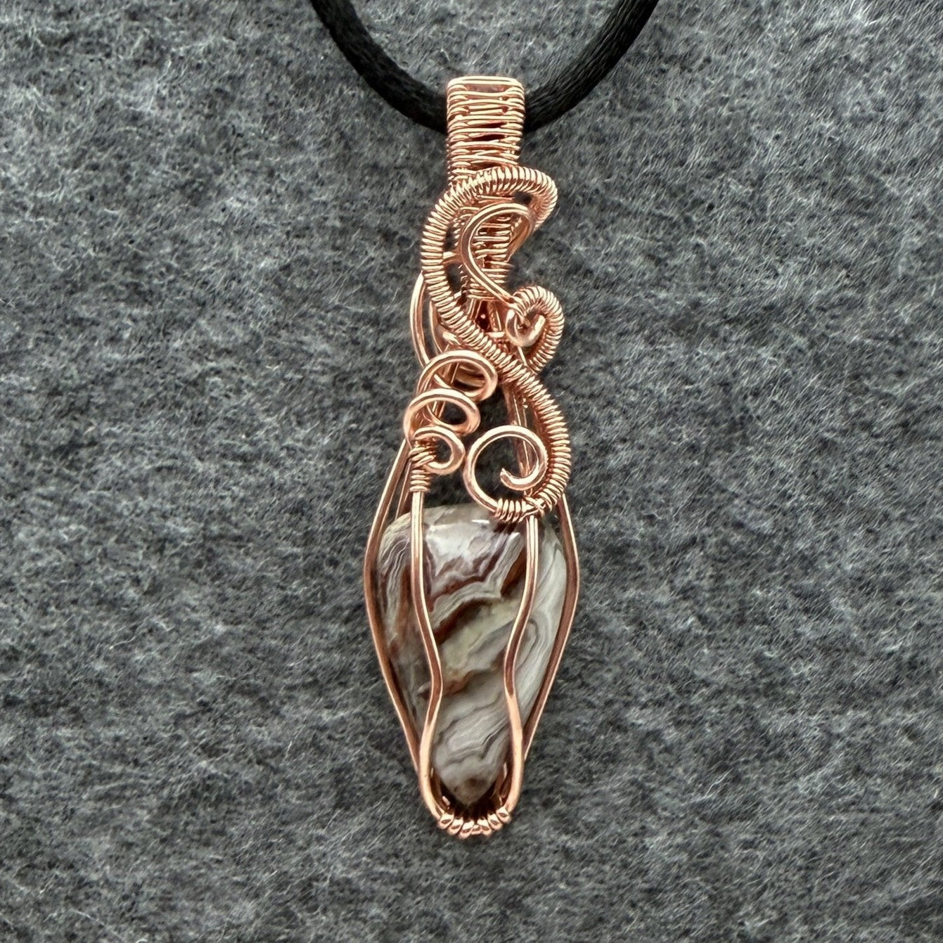 Front view of a handmade wire-wrapped pendant necklace featuring a crazy lace agate stone enclosed in copper wire, suspended from a black cord against a textured gray background.