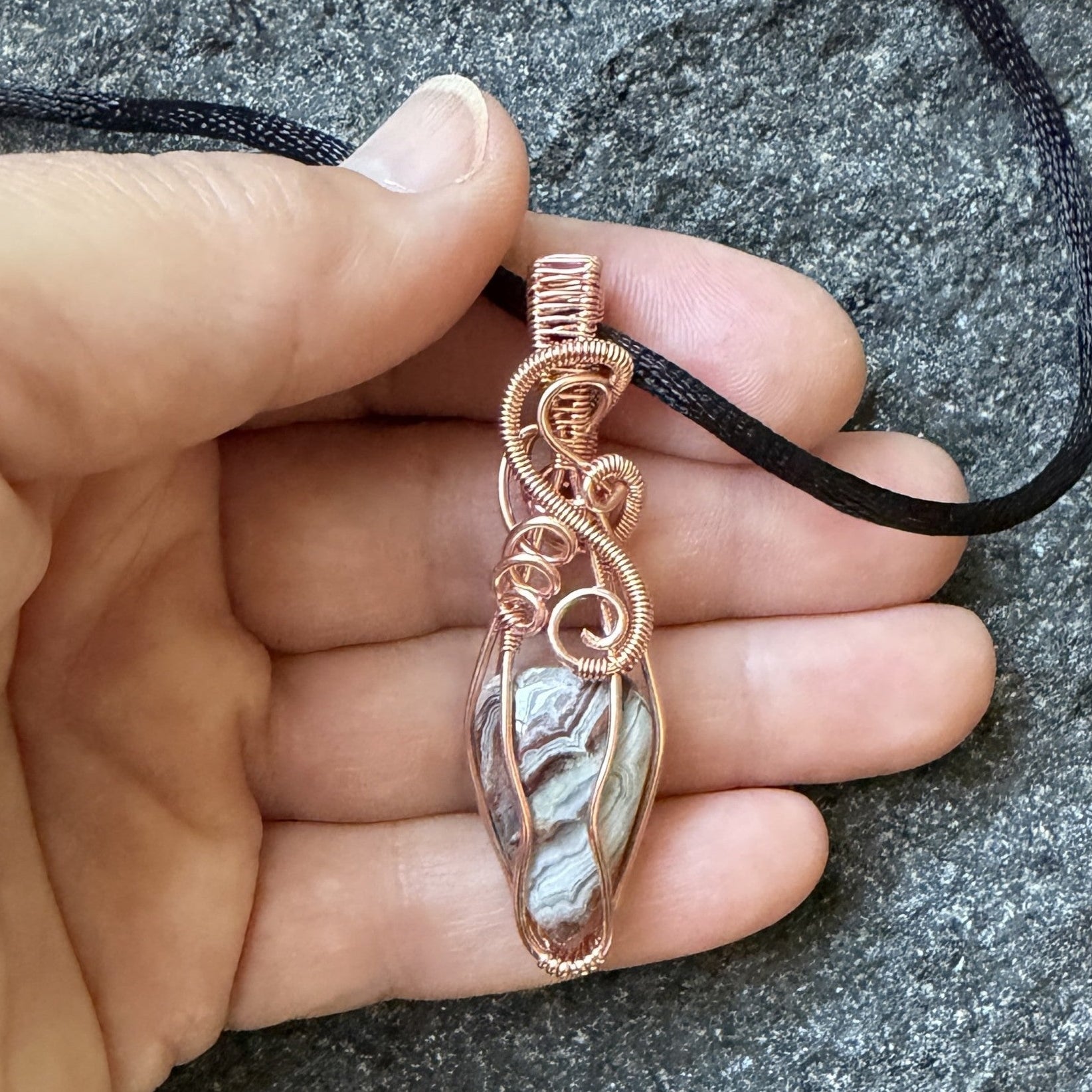 Front view of a handmade wire-wrapped pendant necklace featuring a crazy lace agate stone enclosed in copper wire, suspended from a black cord, held in the artist's hand.