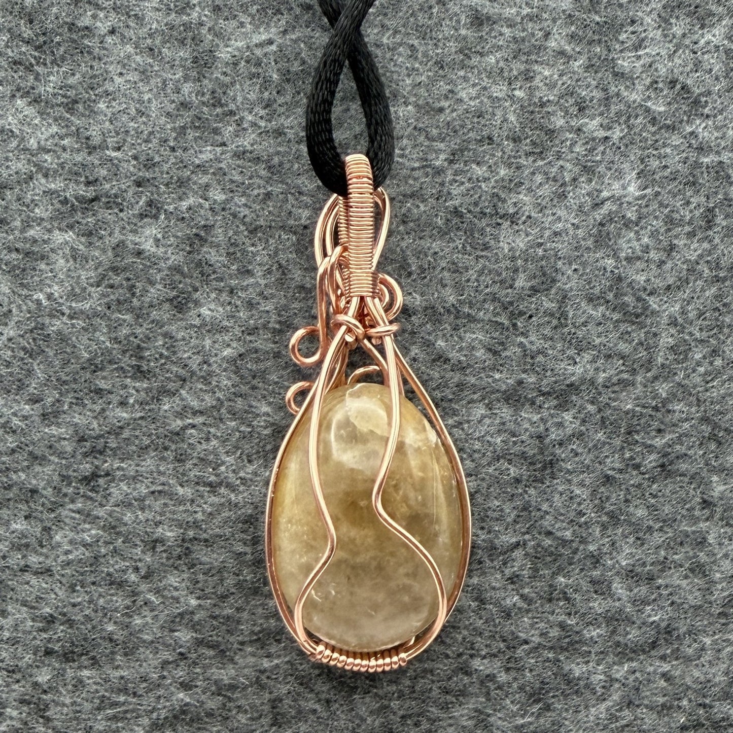 Back view of a teardrop-shaped pendant necklace featuring a pale yellow quartz stone wrapped in copper wire, suspended from a black cord against a textured gray background.