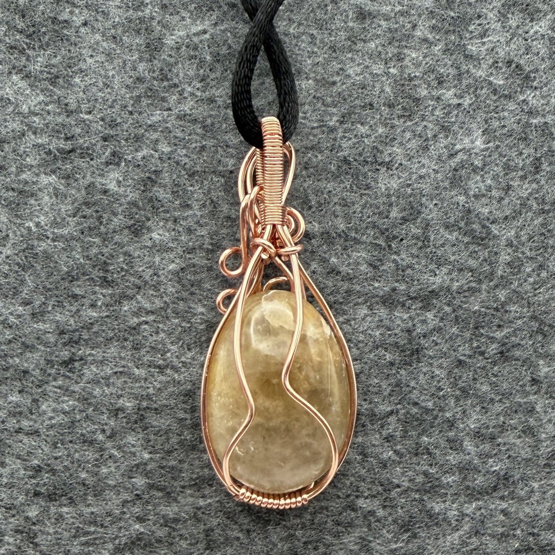 Back view of a teardrop-shaped pendant necklace featuring a pale yellow quartz stone wrapped in copper wire, suspended from a black cord against a textured gray background.