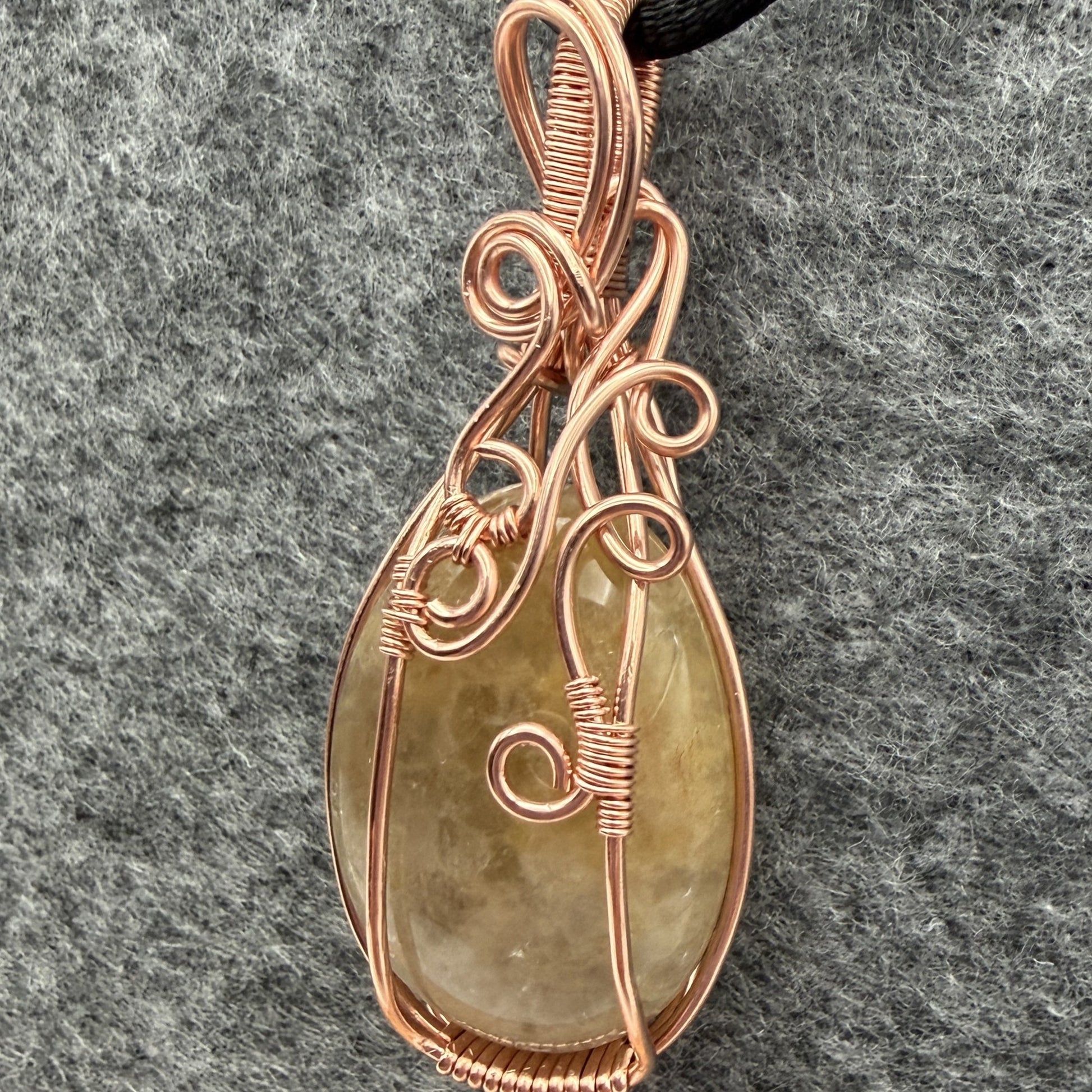 Detail view of a teardrop-shaped pendant necklace featuring a pale yellow quartz stone wrapped in copper wire, suspended from a black cord against a textured gray background.
