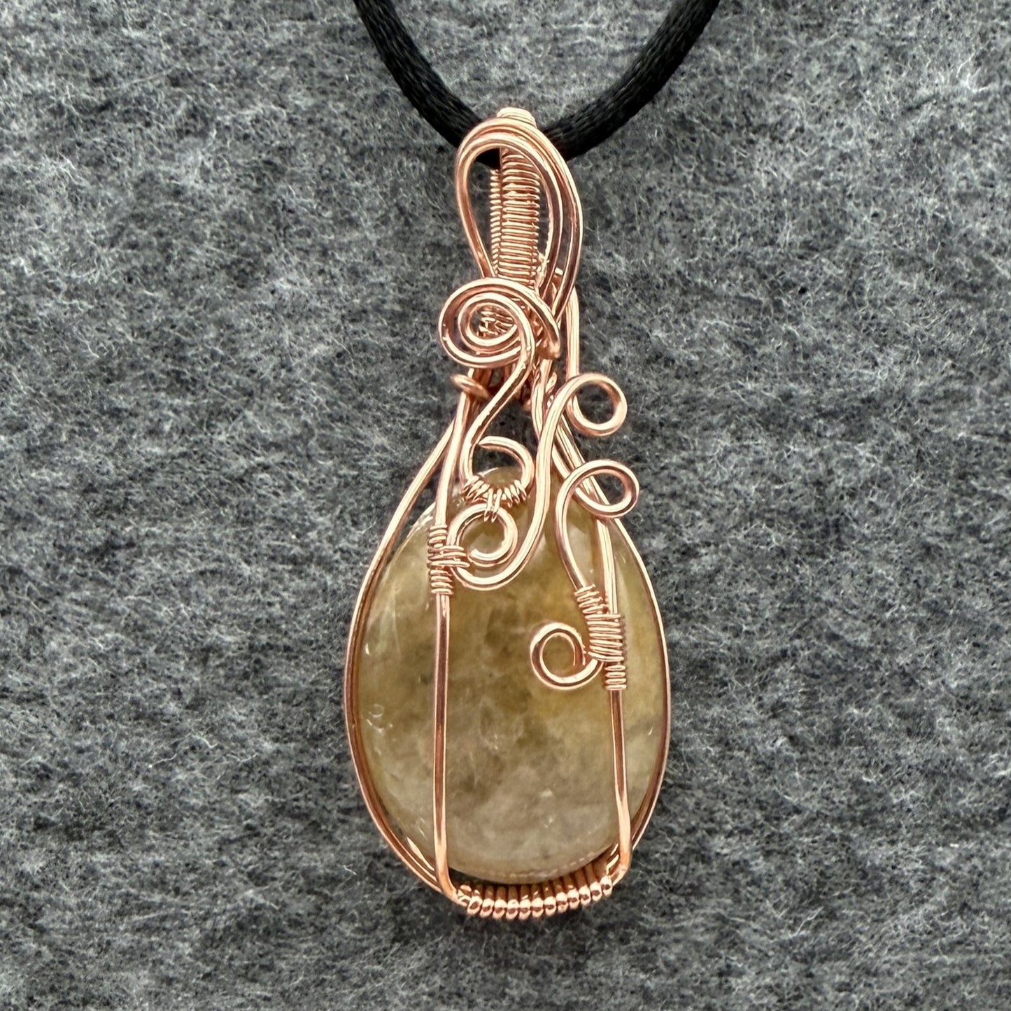 Front view of a teardrop-shaped pendant necklace featuring a pale yellow quartz stone wrapped in copper wire, suspended from a black cord against a textured gray background.