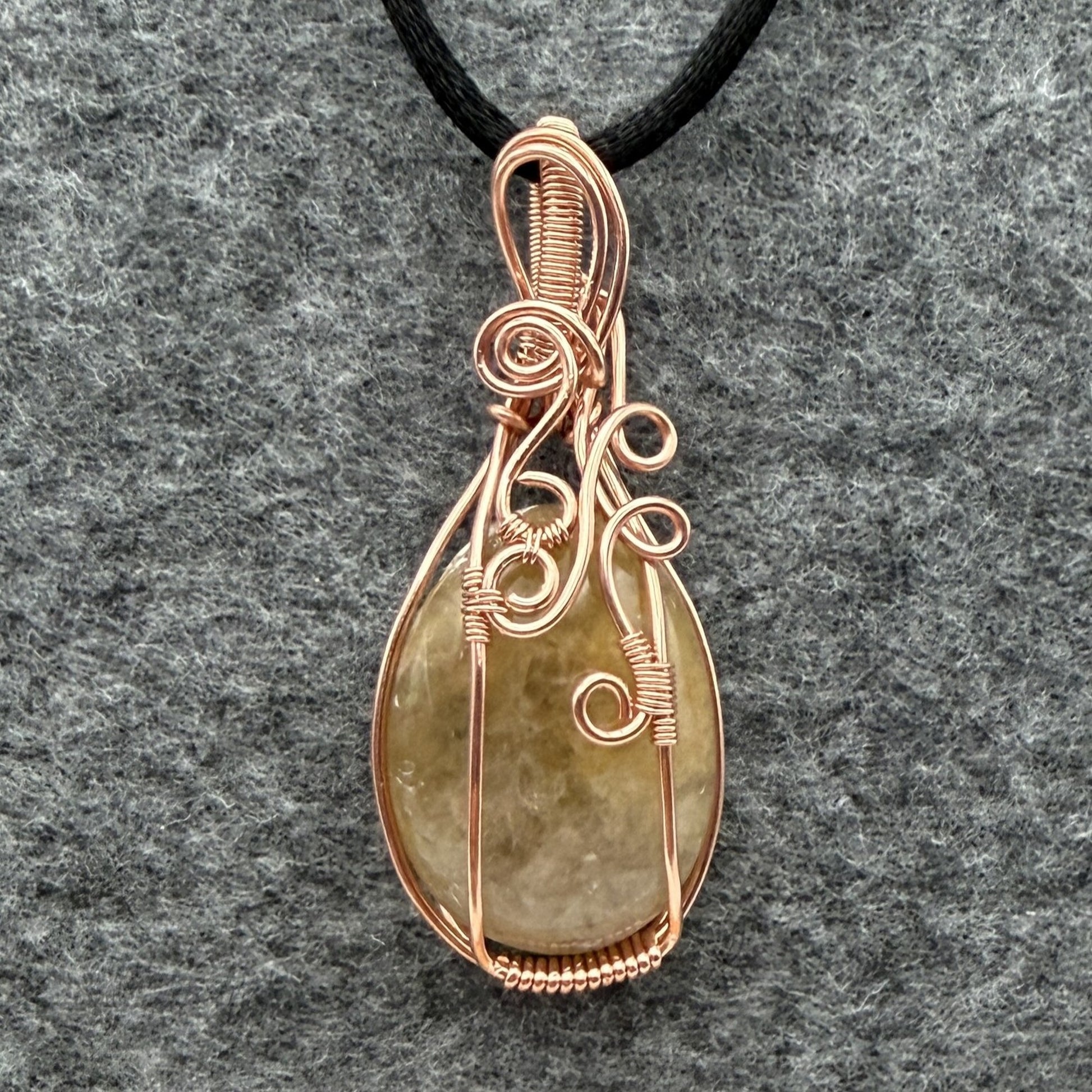 Front view of a teardrop-shaped pendant necklace featuring a pale yellow quartz stone wrapped in copper wire, suspended from a black cord against a textured gray background.