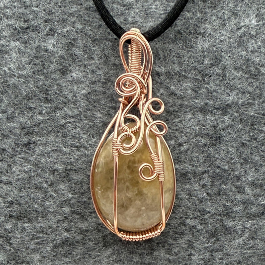 Front view of a teardrop-shaped pendant necklace featuring a pale yellow quartz stone wrapped in copper wire, suspended from a black cord against a textured gray background.