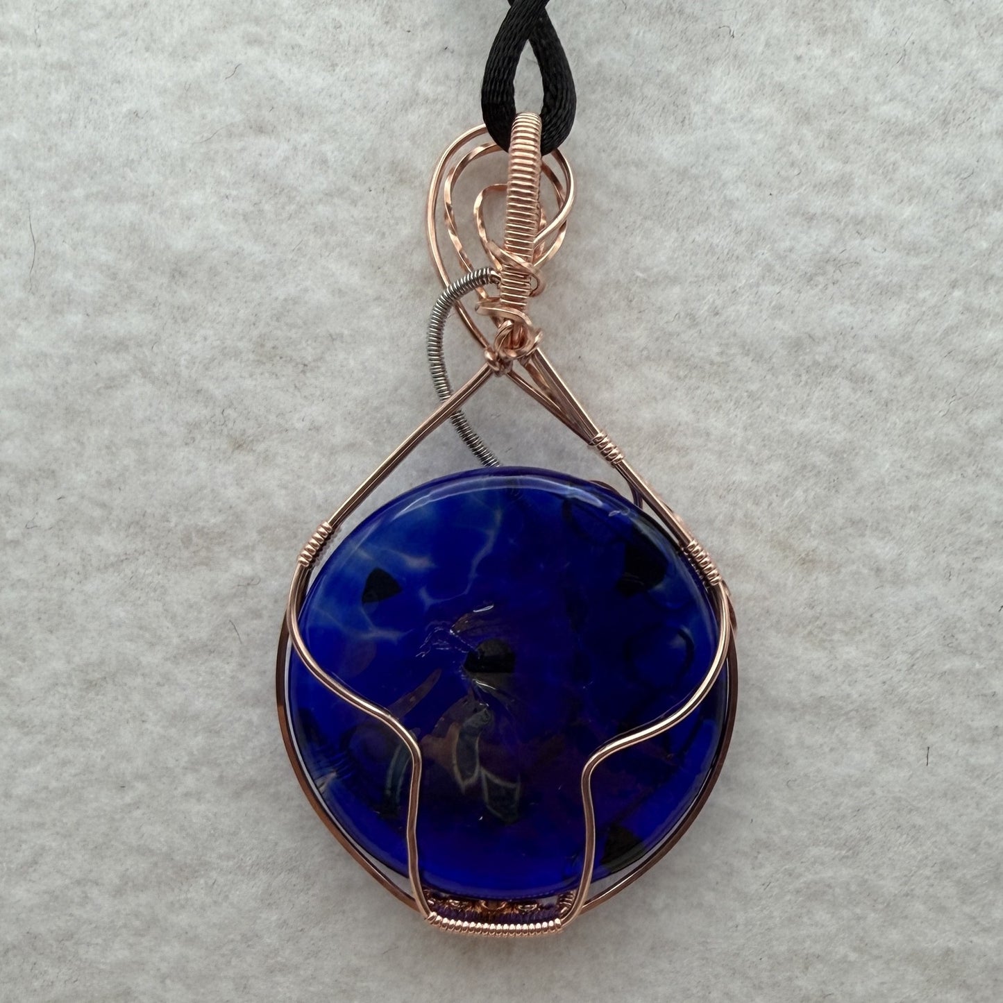 Back view of a teardrop-shaped, cobalt blue glass pendant necklace wrapped in copper and stainless steel wire, hanging from a black cord on a textured white background.
