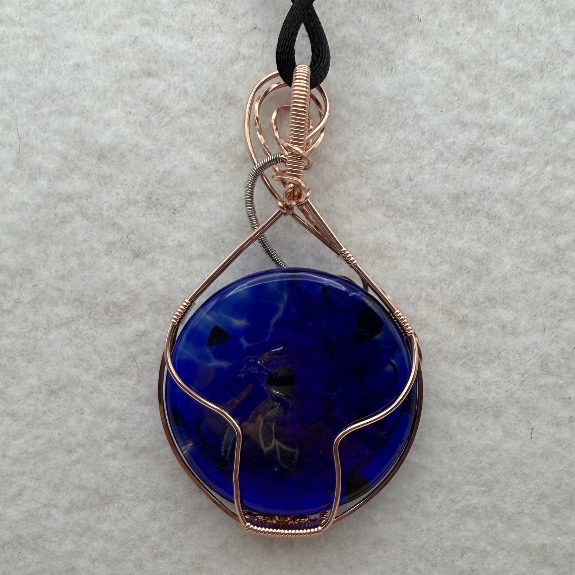 Back view of a teardrop-shaped, cobalt blue glass pendant necklace wrapped in copper and stainless steel wire, hanging from a black cord on a textured white background.