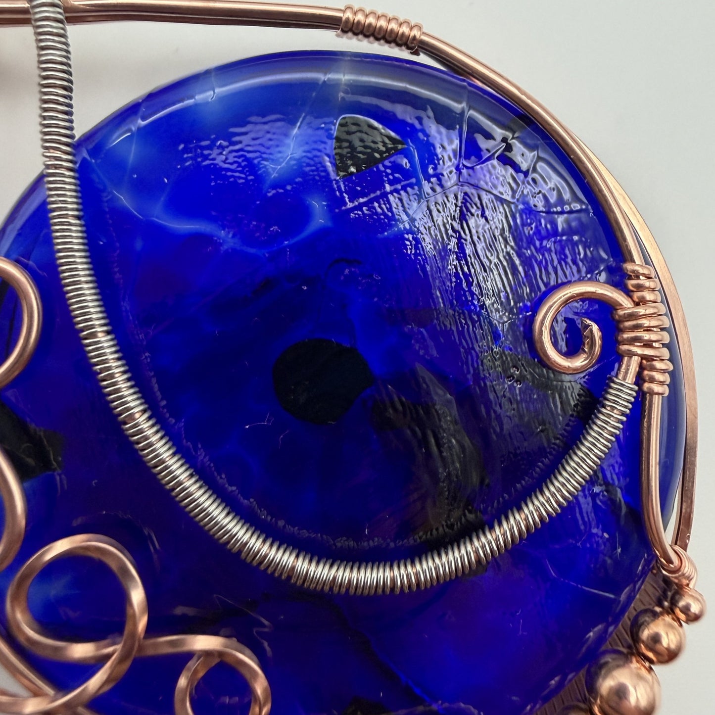Detail view of a teardrop-shaped, cobalt blue glass pendant necklace wrapped in copper and stainless steel wire, hanging from a black cord on a textured white background.
