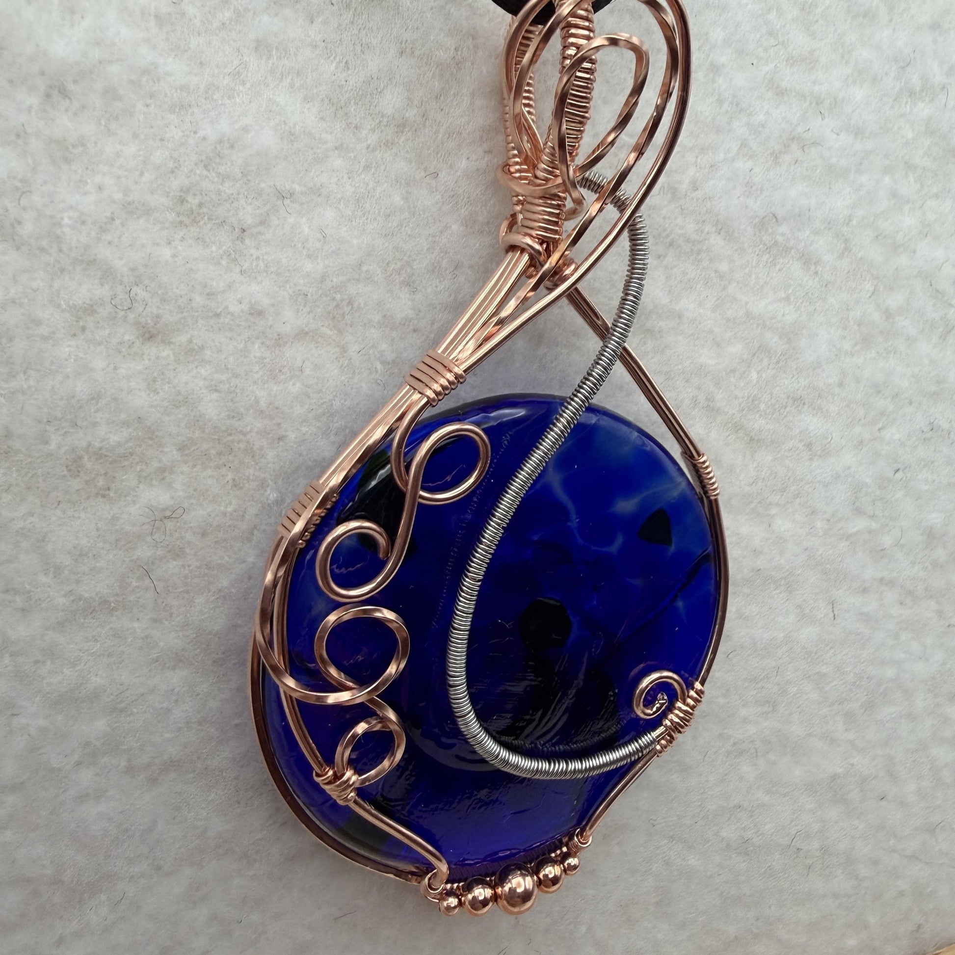 Detail view of a teardrop-shaped, cobalt blue glass pendant necklace wrapped in copper and stainless steel wire, hanging from a black cord on a textured white background.