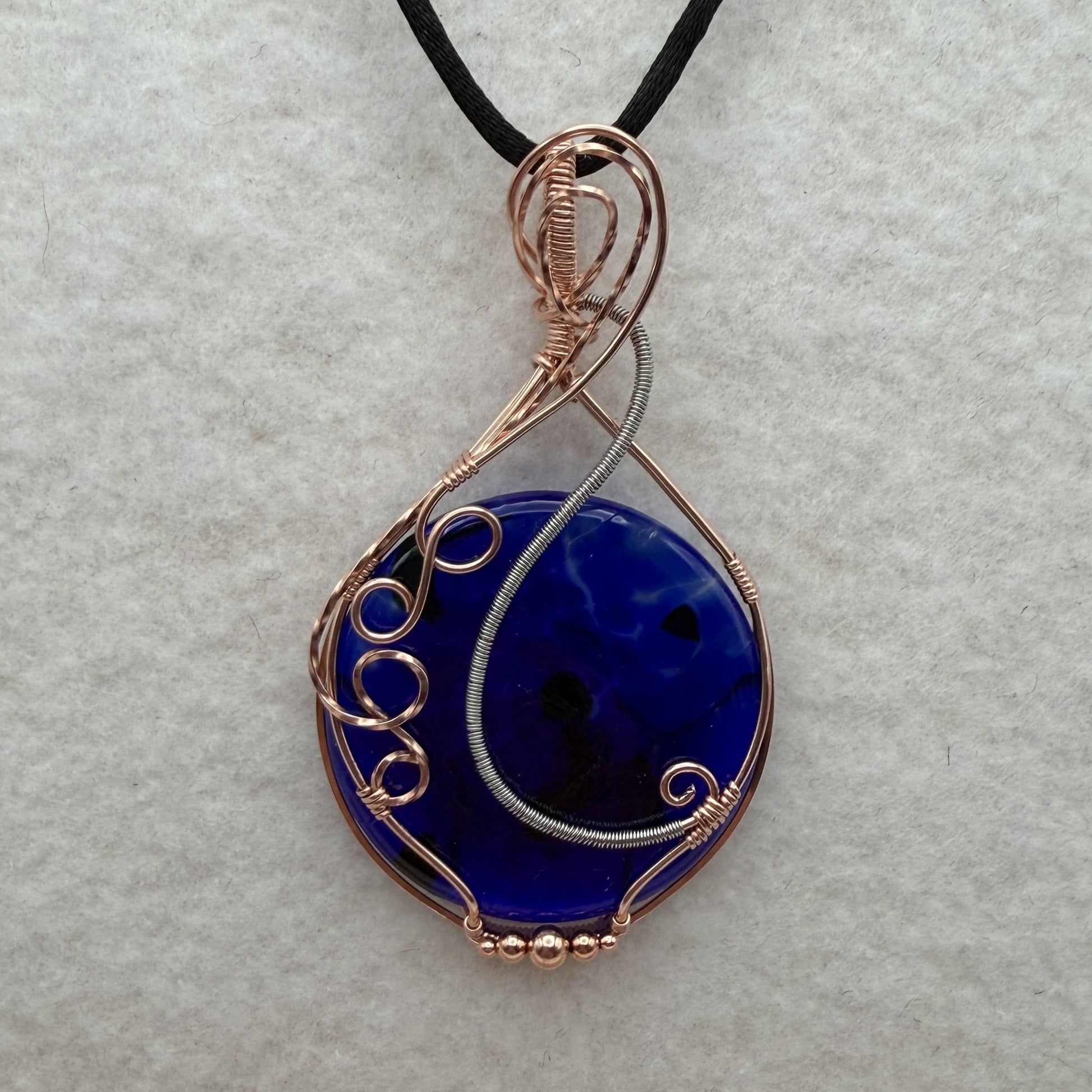 Front view of a teardrop-shaped, cobalt blue glass pendant necklace wrapped in copper and stainless steel wire, hanging from a black cord on a textured white background.