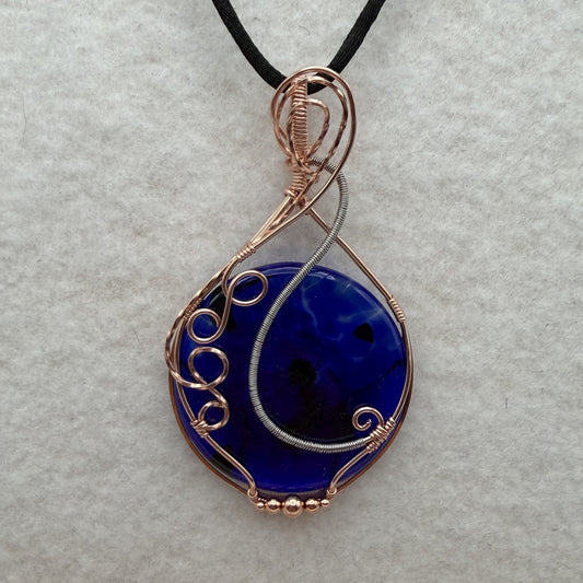 Front view of a teardrop-shaped, cobalt blue glass pendant necklace wrapped in copper and stainless steel wire, hanging from a black cord on a textured white background.